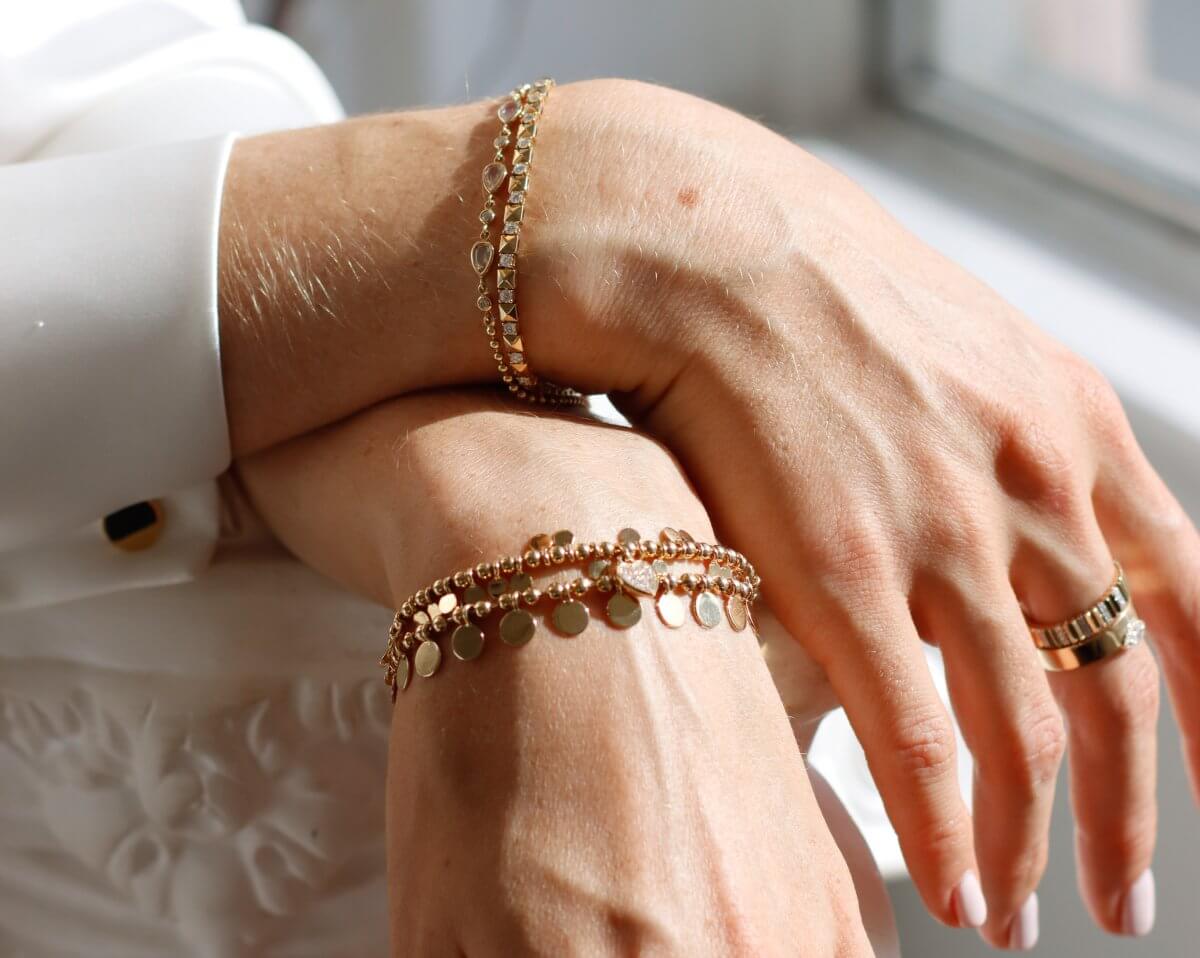 Close-up of two hands wearing gold bracelets and a gold ring, resting on a white surface. The bracelets have small circular charms and beads, and the person is wearing a white shirt.