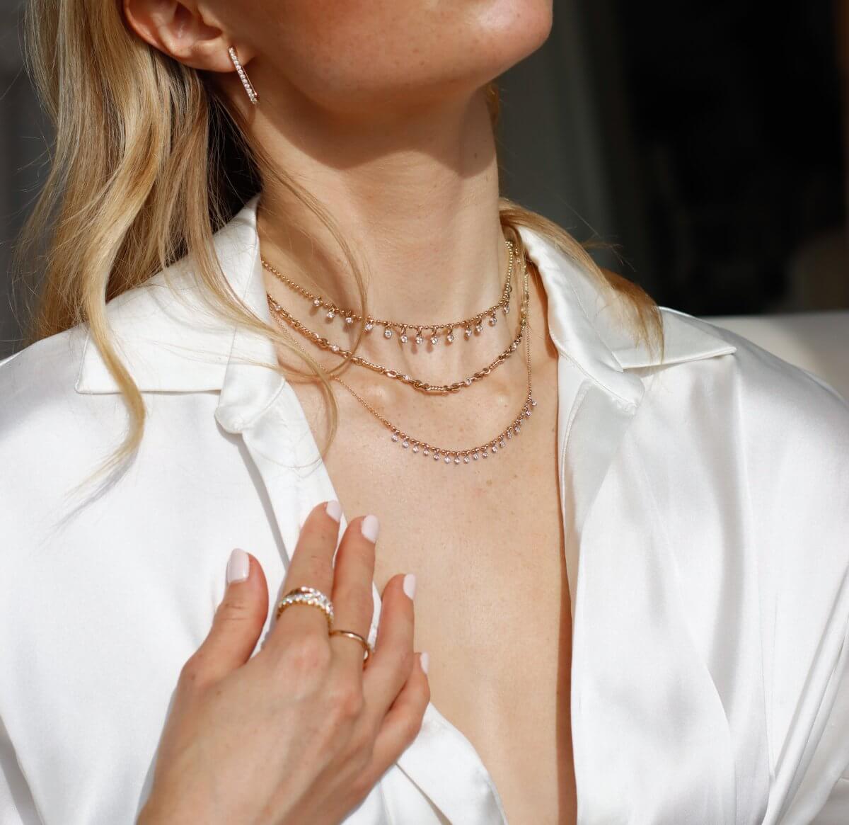 Person wearing a white blouse, layered gold necklaces with small gems, a gold ring, and an earring. Their hand is touching their blouse near the neck. The focus is on the jewelry.