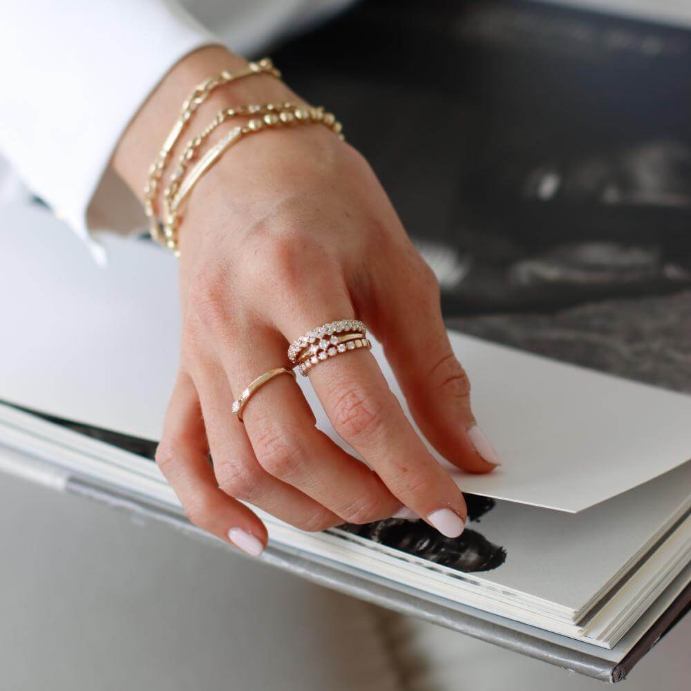 A hand adorned with gold bracelets and diamond rings rests on an open book. The person wears a white sleeve, and the nails are manicured in a light color.