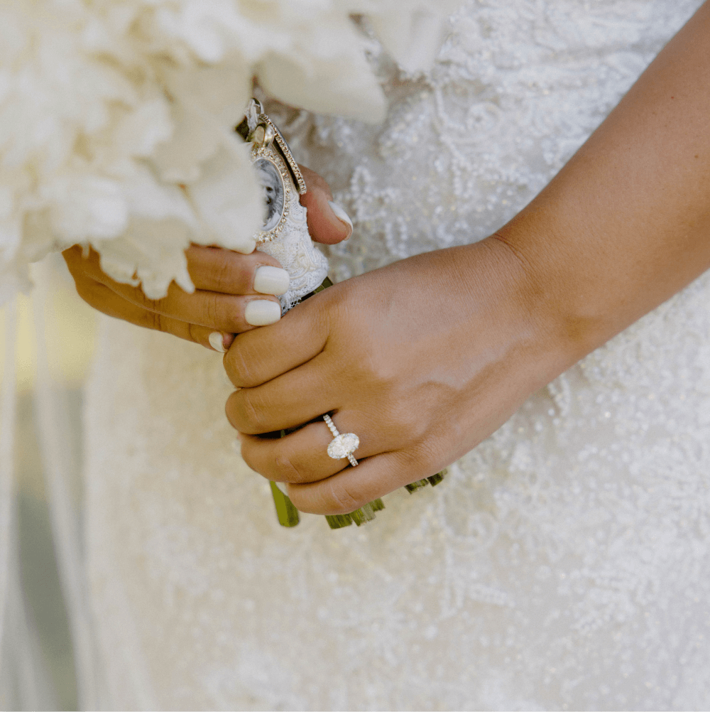 A bride in a beaded lace gown holds a bouquet wrapped with a sentimental item. Her manicured hand features a sparkling engagement ring.