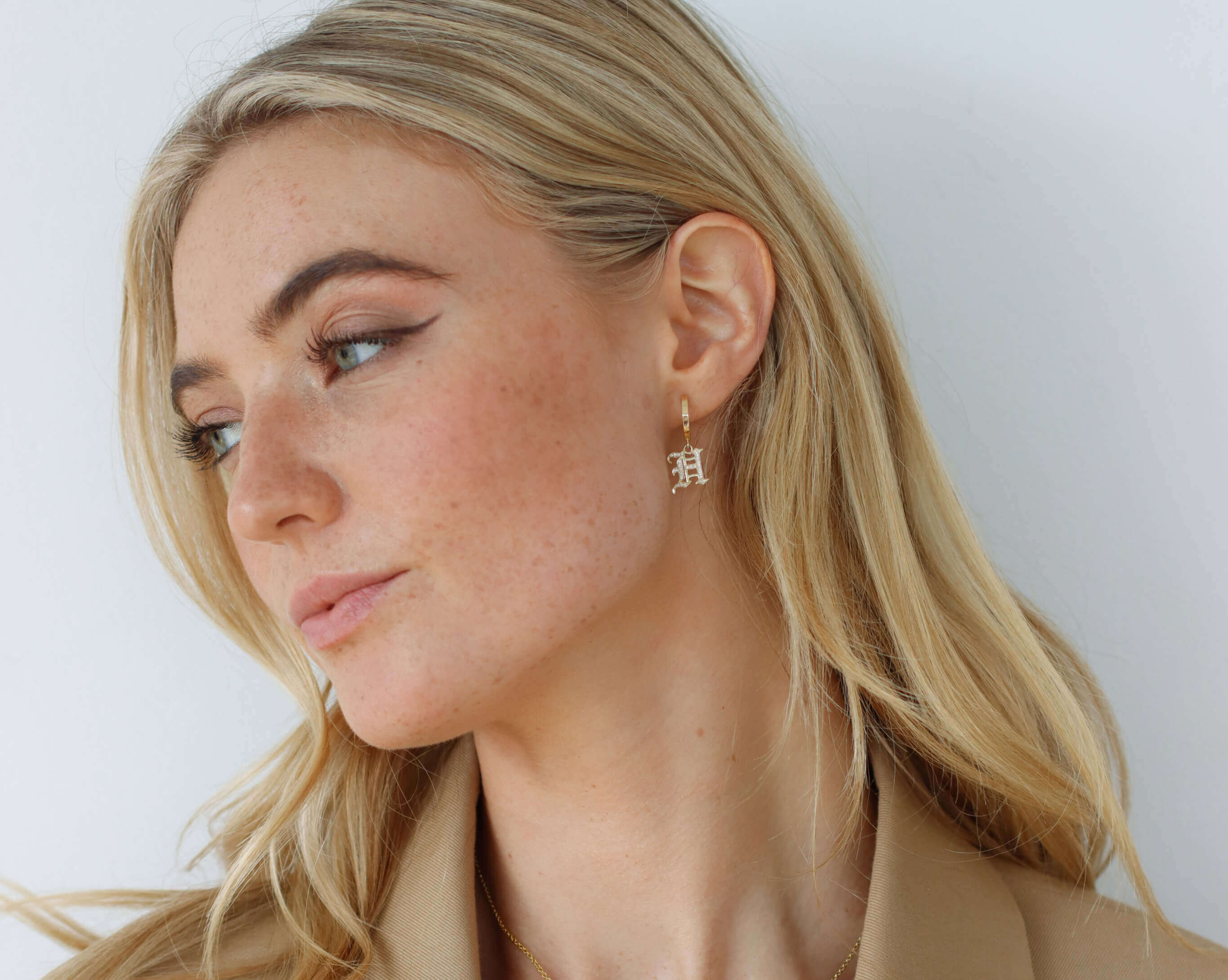 A person with long blonde hair is looking to the side. They are wearing a beige blazer and gold hoop earrings with a pendant. The background is a plain, light-colored wall.