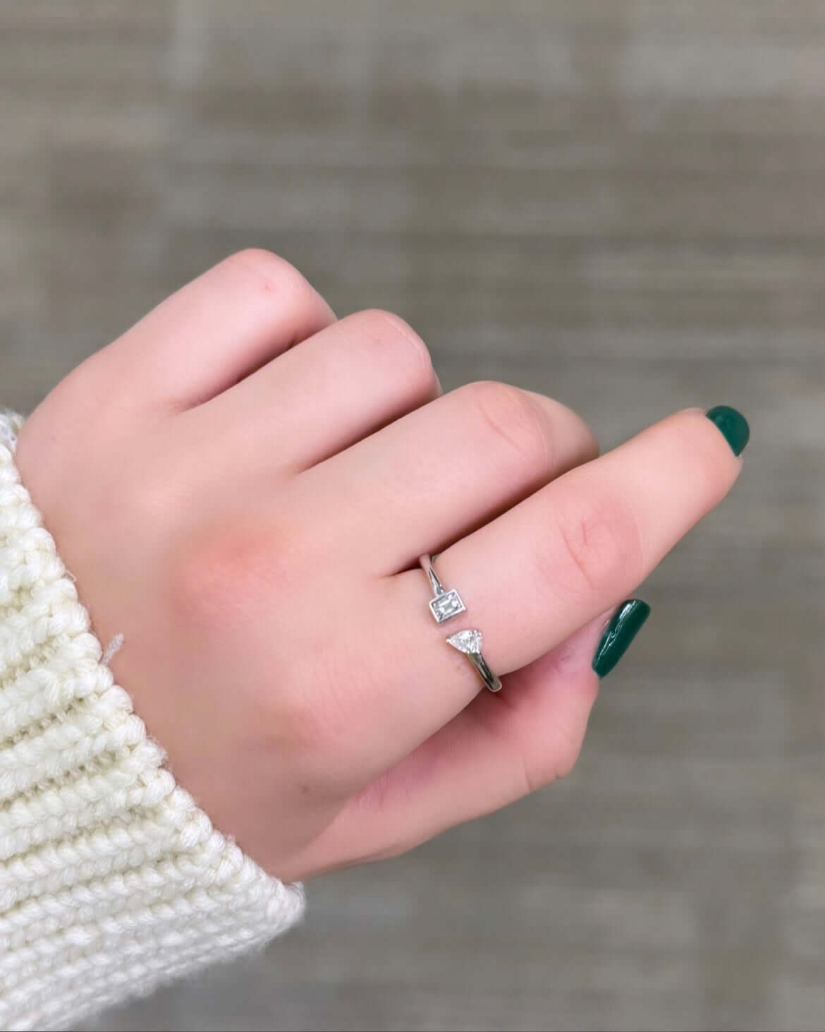 A hand with green polish showcases the Emerald & Pear Toi Et Moi Ring, symbolizing love with dual gemstones. The person wears a white knitted sweater against an earthy-toned background. This captivating piece is available for preorder.
