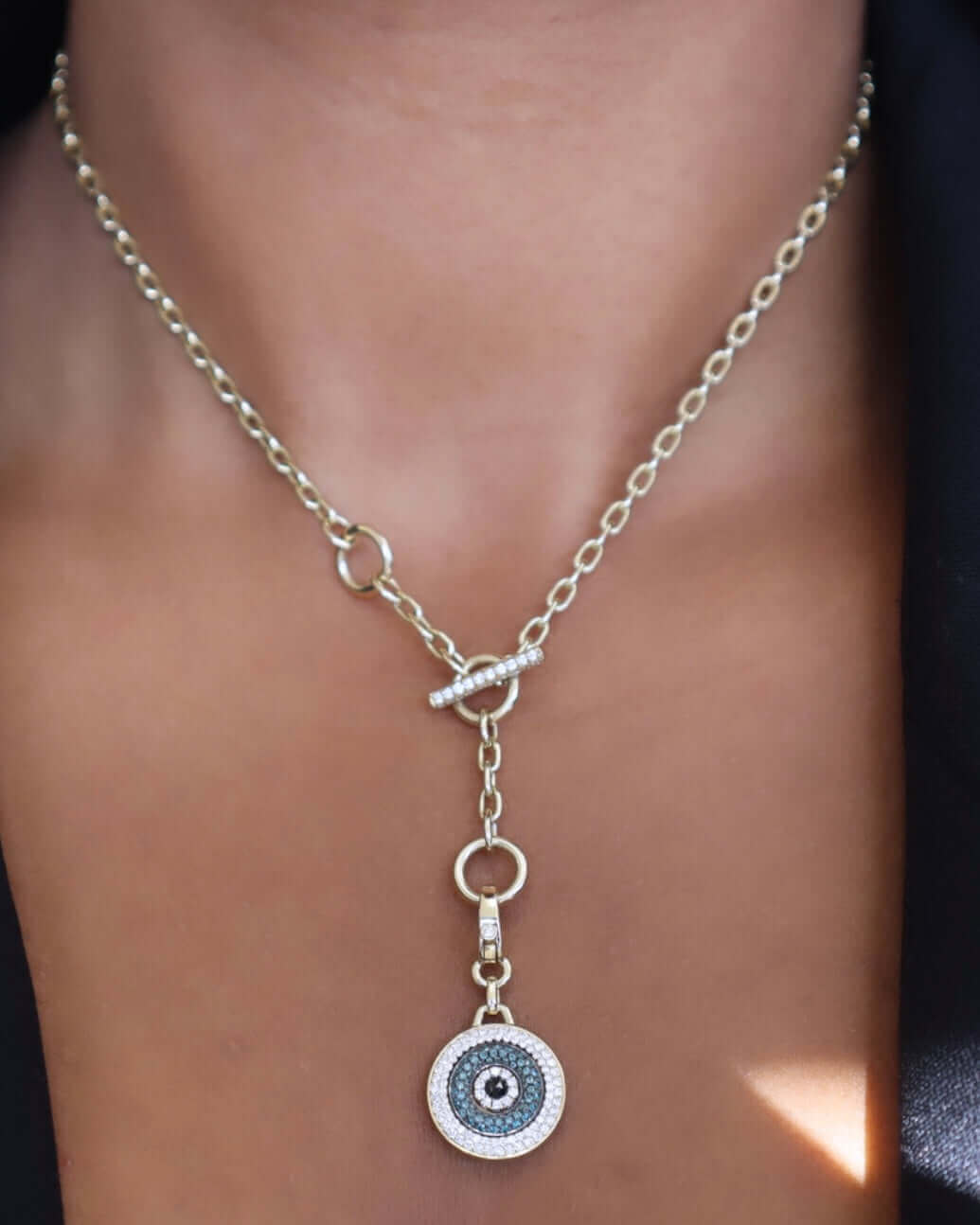 A woman accessorizes her black top with a gold necklace from Janet Heller Fine Jewelry, highlighting a Round Blue Diamond Evil Eye Charm. The custom Y-shaped necklace features an eye pendant with concentric circles in black, blue, and white and is secured by a toggle clasp.