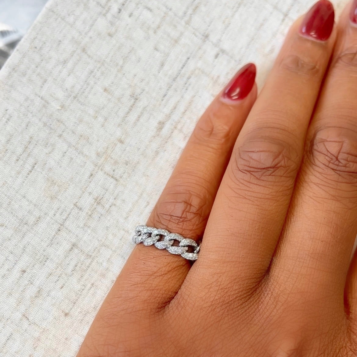 A hand with red nail polish wearing a Small Diamond Chain Ring from Janet Heller Fine Jewelry rests on a textured surface.