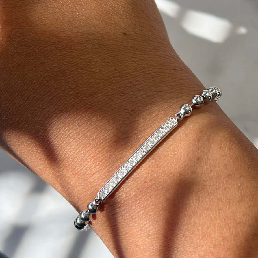 A wrist is adorned with the Toilet Chain Diamond ID Bracelet, a silver bracelet from Janet Heller Fine Jewelry featuring a central bar encrusted with tiny sparkling stones, complemented by small round silver beads on both sides, set against blurred shadow patterns.