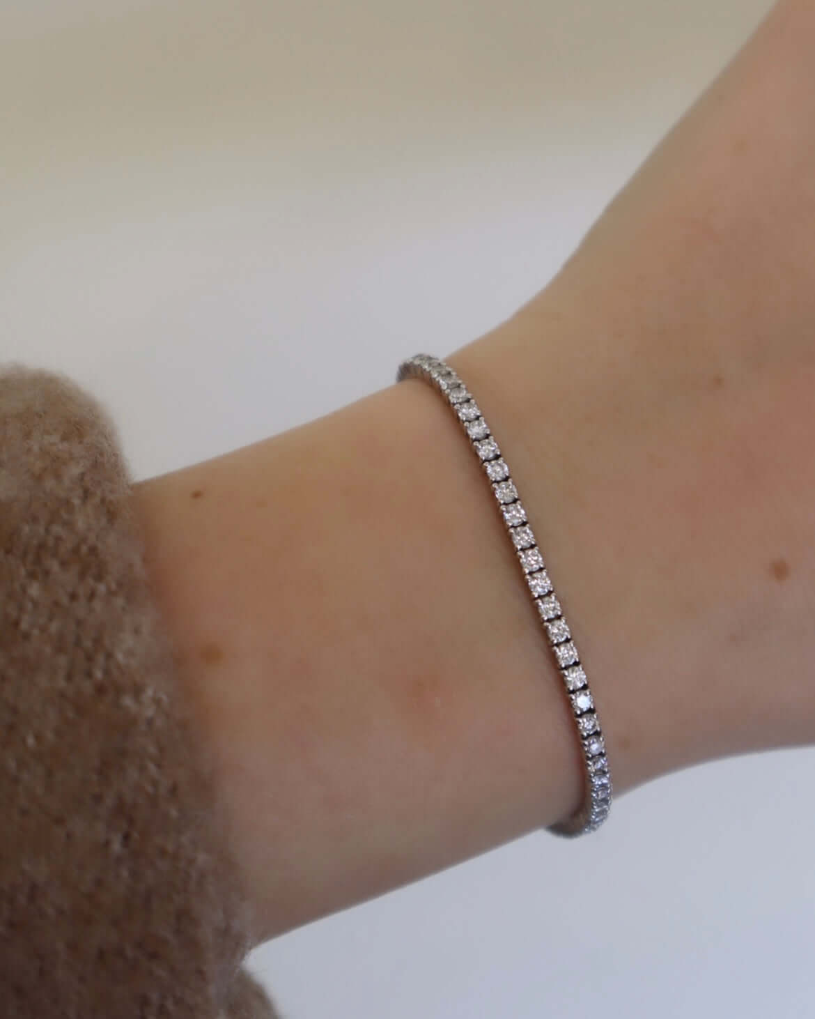 A close-up shows a wrist wearing a 2ct Round Diamond Tennis Bracelet in silver with lab-grown diamonds, paired beautifully with a brown fuzzy sweater against a neutral backdrop.