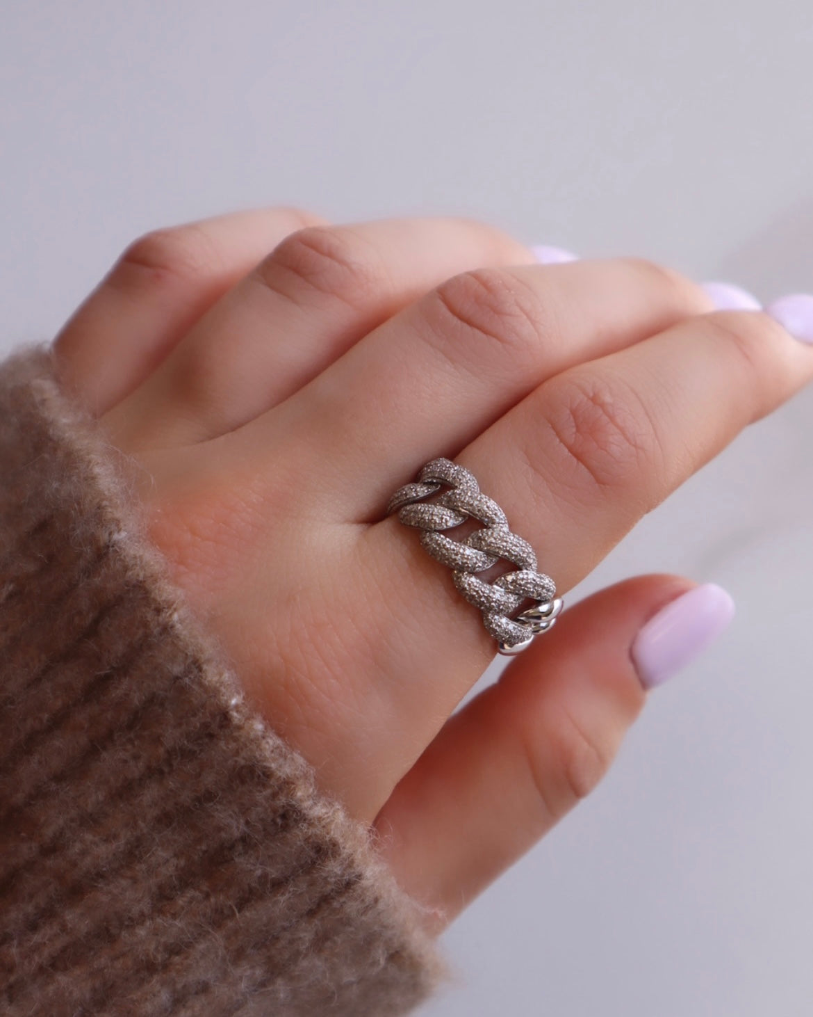 A light-skinned hand features the Diamond Hunter Ring, a chunky silver chain-link piece from Janet Hellers fine jewelry collection with a textured design. The nails are painted light lavender, and the wearer is clad in a cozy brown ribbed sweater against a softly blurred background.