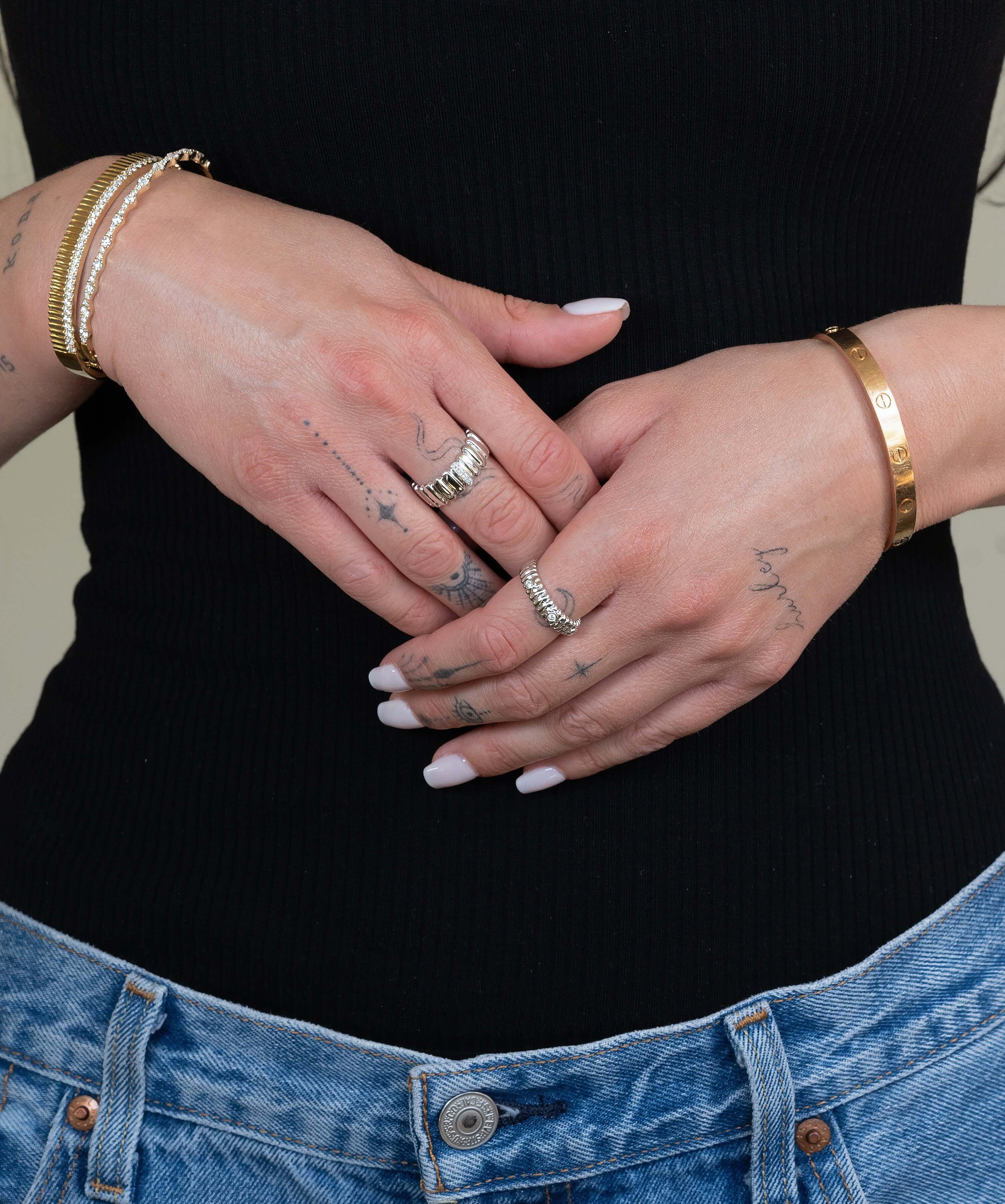 A person in a black ribbed top and blue jeans stands with crossed hands, wearing gold bracelets and the Diamond Ridge Rio Ring as fine jewelry. Their white-painted nails enhance the silver rings, while tattoos adorn their hands and arms.