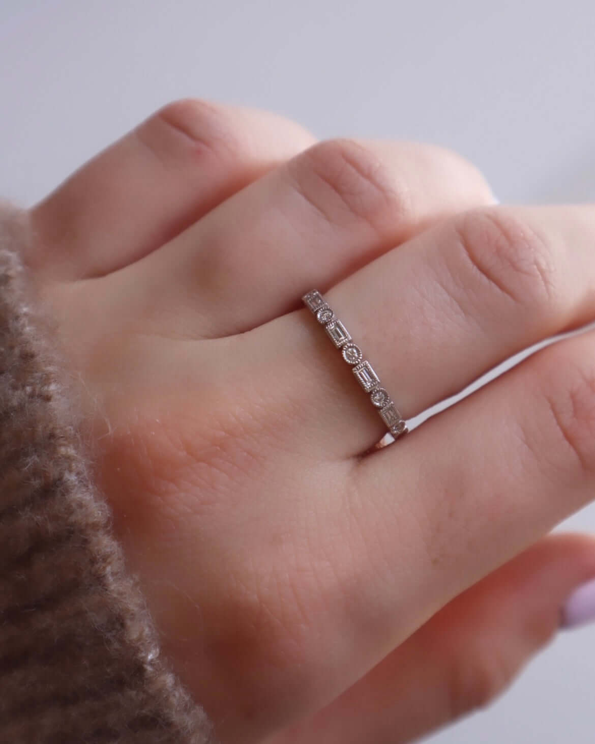 A hand displays the Diamond Shapes Ring—a delicate silver piece adorned with small, evenly spaced gems from the Duplicate SKU collection (RIN-119-5-W, $573.00) by Janet Heller. The hand rests on a soft, fuzzy brown sleeve against a softly blurred background. Perfect as a preorder choice for those seeking timeless elegance.
