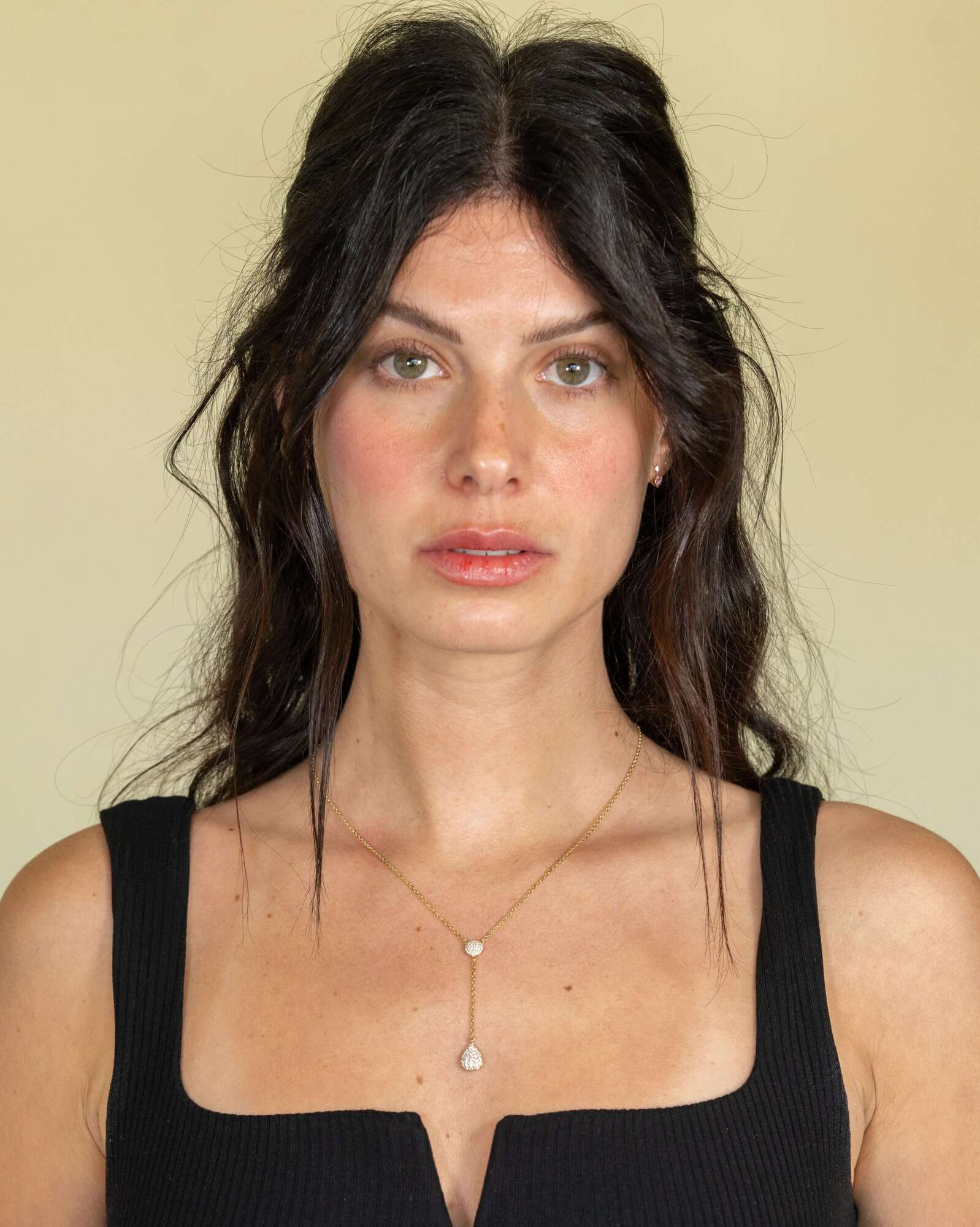 A woman with long, wavy dark hair and green eyes, in a black sleeveless top, looks at the camera. She wears a Janet Heller Fine Jewelry Diamond Teardrop & Round Lariat Necklace. The background is plain beige.