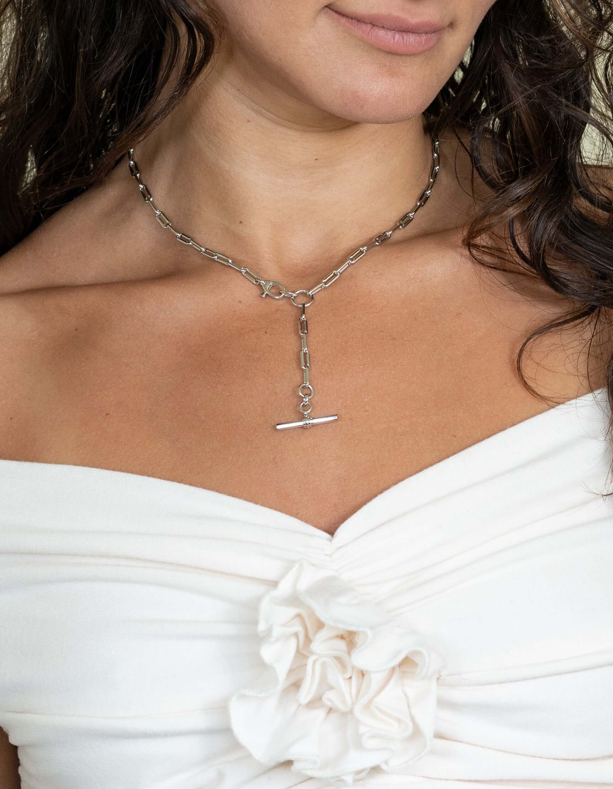 They highlight the Toggle Link Kristen Necklace, a striking silver chain with a T-bar pendant, over an off-the-shoulder white top adorned with floral details. Their wavy hair adds elegance, emphasizing the custom touches of their stylish ensemble.