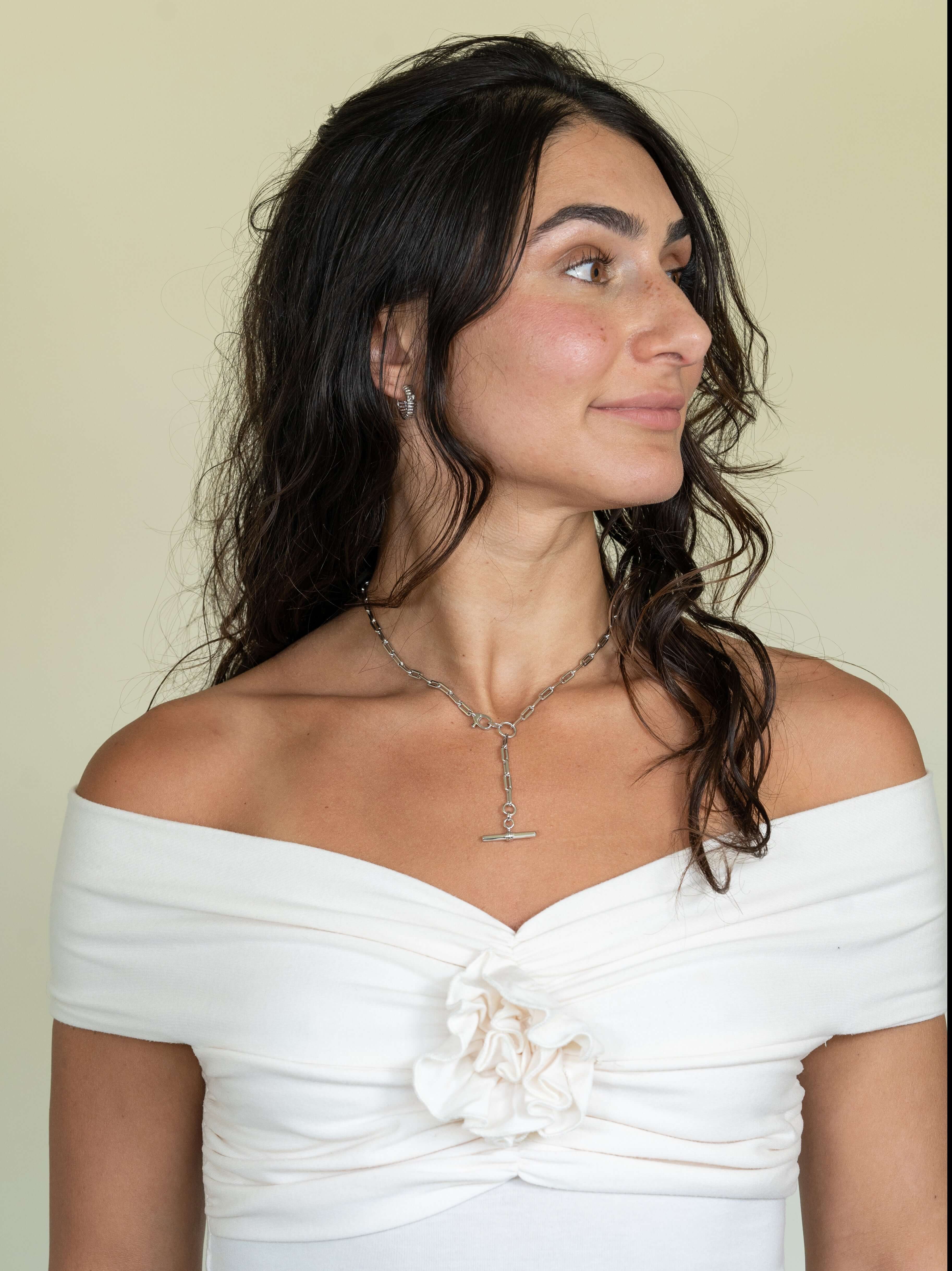 A woman with long, wavy dark hair looks left, showcasing a white off-the-shoulder top with floral details. Her look is completed by the stunning Toggle Link Kristen Necklace—a silver piece that sparkles against the plain, light background.