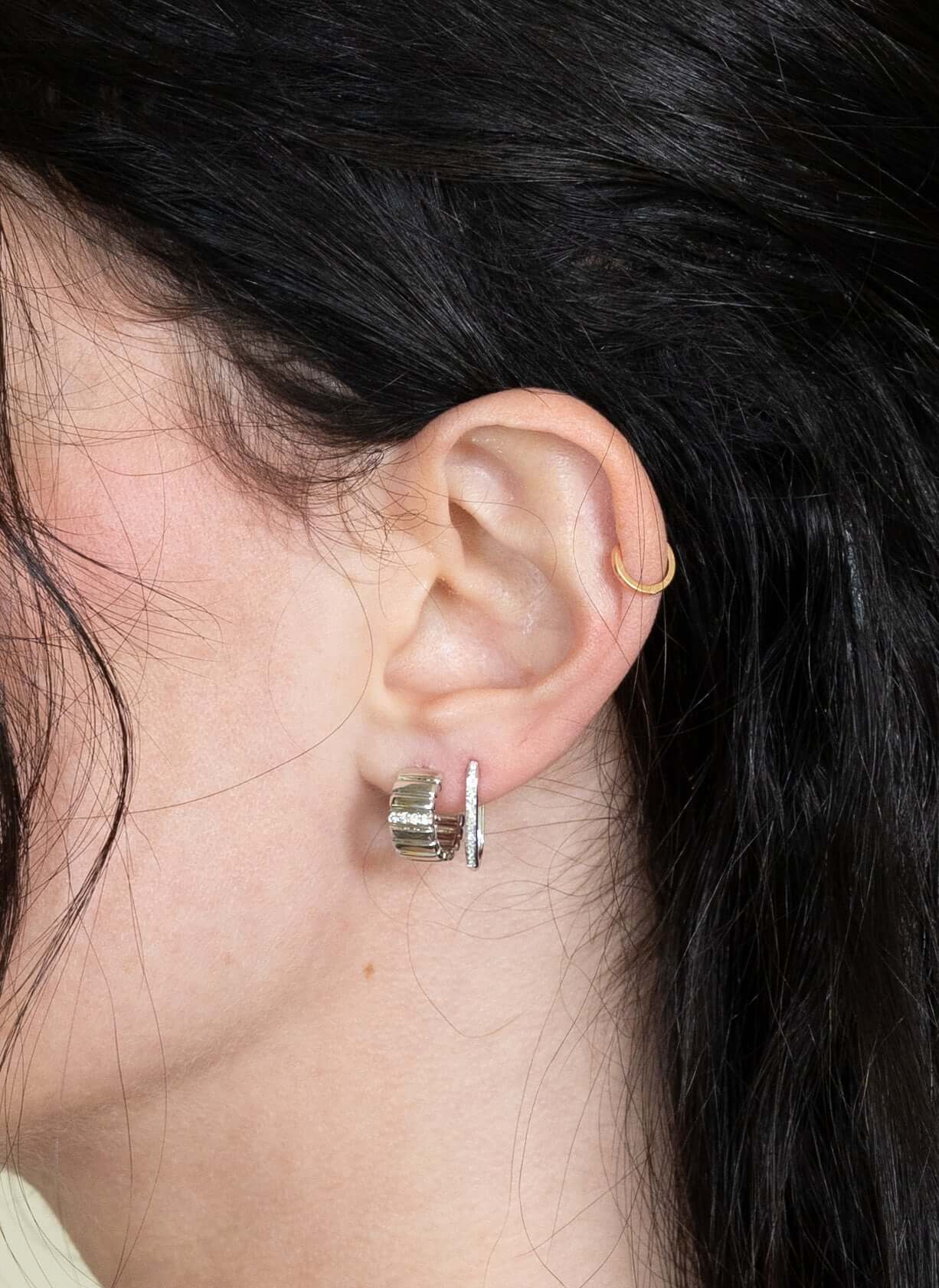 A close-up shows a persons ear with dark hair, adorned with the Diamond Ridge Rio Huggies in silver on the earlobe and a sleek gold hoop on the cartilage. The neutral background accentuates these exquisite jewelry pieces.