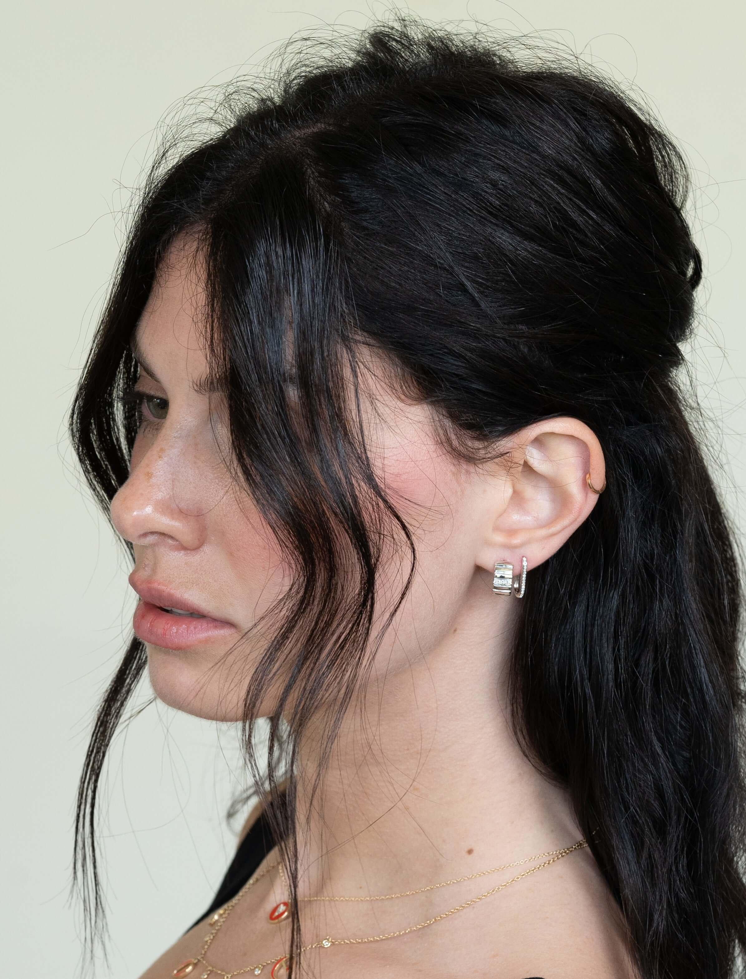 A person with long black hair and hoop earrings, showcased in profile against a neutral backdrop, wears Diamond Ridge Rio Huggies. They pair these exquisite earrings with a delicate chain necklace and a black top, exuding elegance.