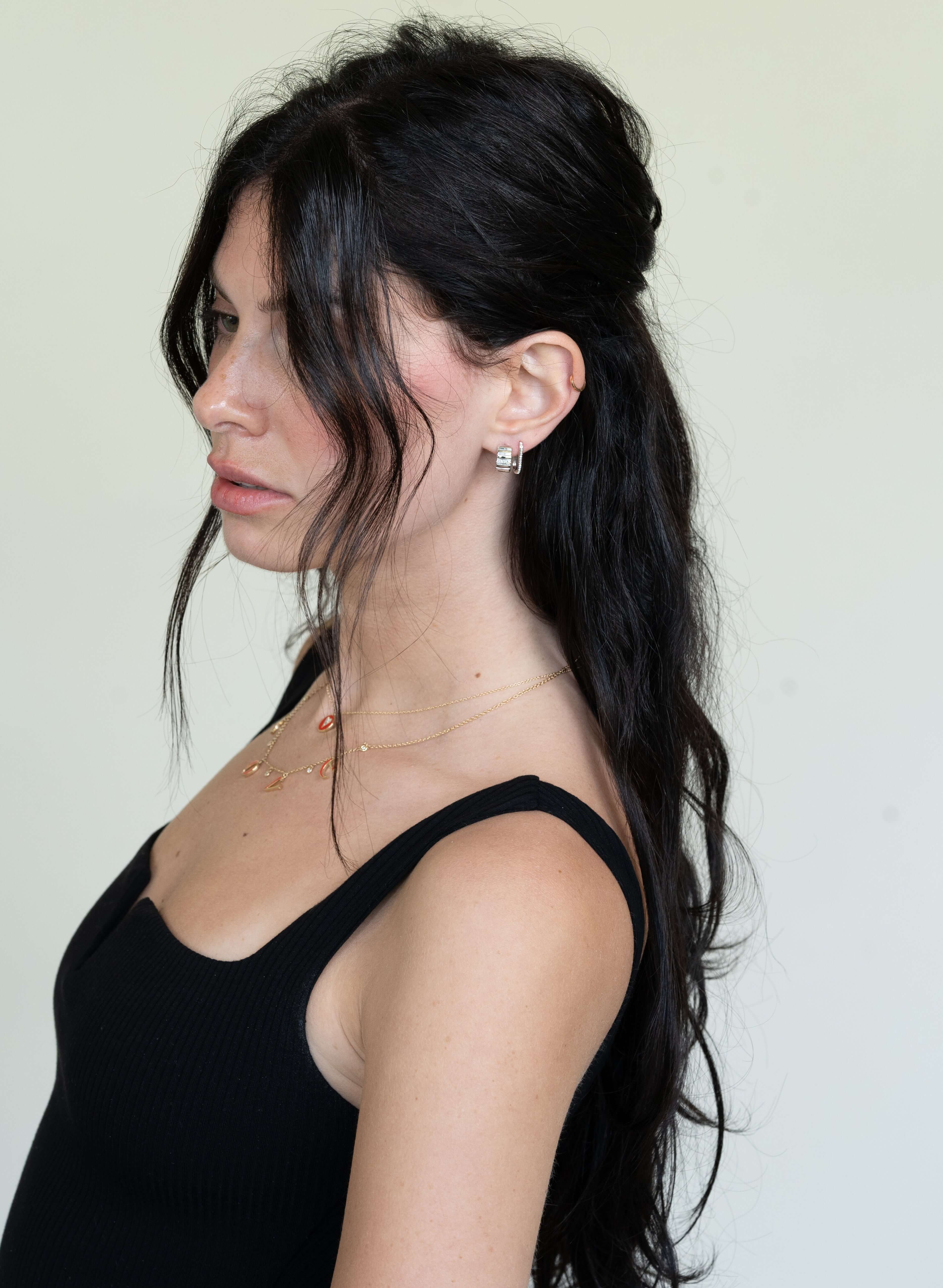 A woman with long, wavy black hair stands profile against a light background. She wears a black sleeveless top, showcasing her fine jewelry—a gold necklace and Diamond Ridge Rio Huggies. Her hair is styled half-up, and she gazes downward with a neutral expression.