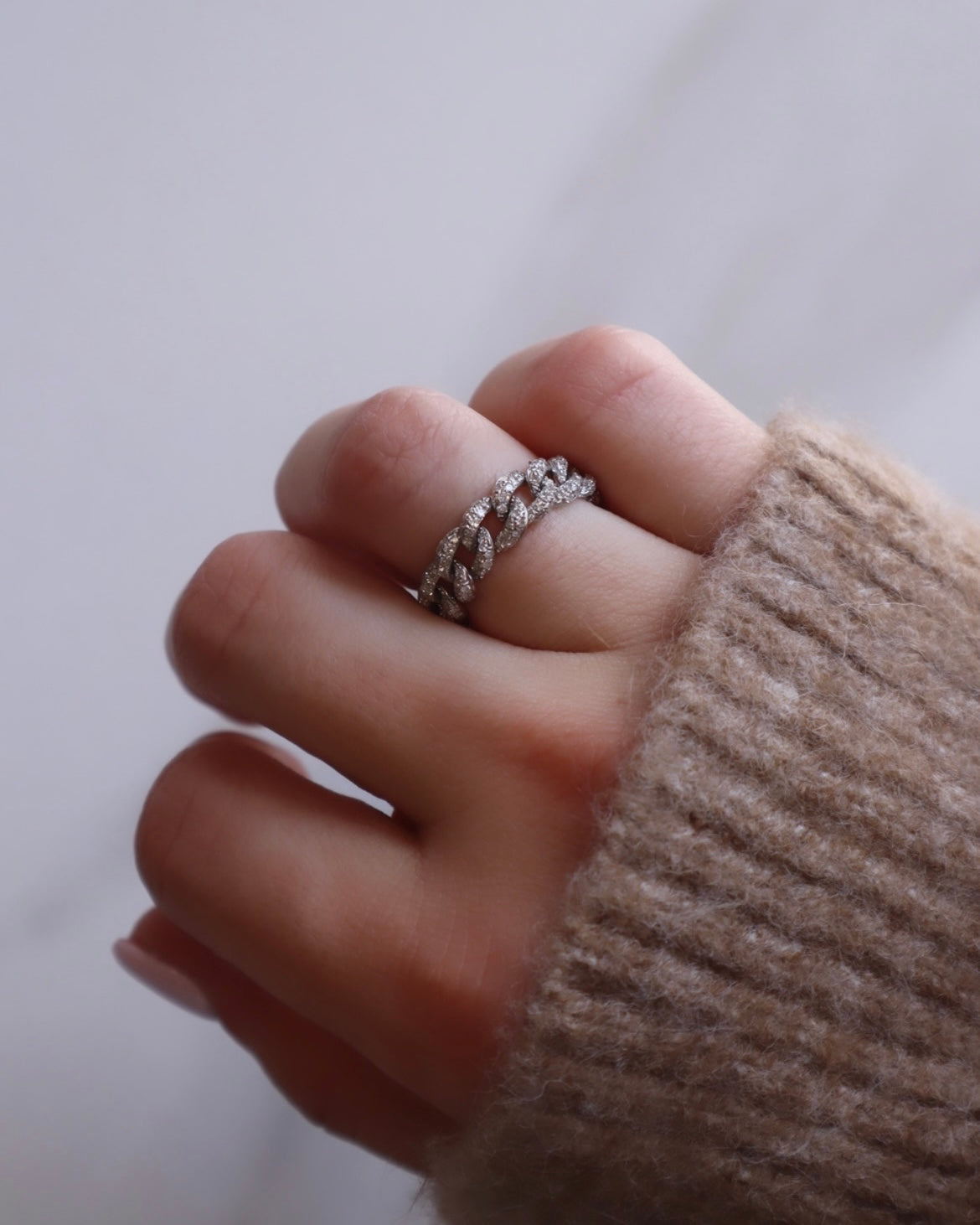 A light-skinned hand wearing a beige sweater displays the Small Diamond Chain Ring by Janet Heller Fine Jewelry. The silver chain-link ring is adorned with sparkling stones and showcased against a soft, blurred white background. Not available in size online; preorder only.