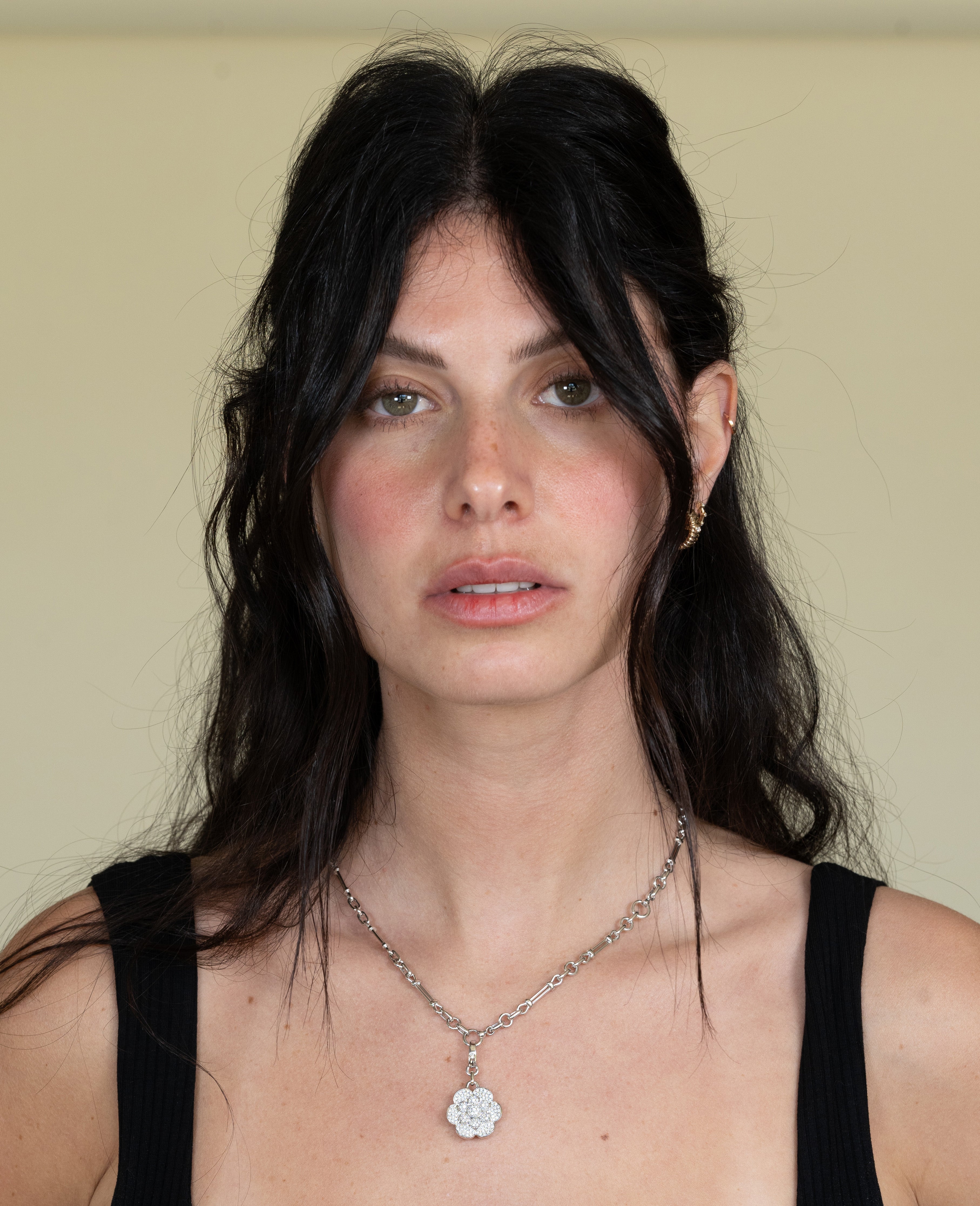 A person with long, dark hair and a neutral expression looks forward, wearing a black top. They have on the Lisbon Chain from Janet Heller Fine Jewelry with charms, and an earring. The background is a plain, light color.
