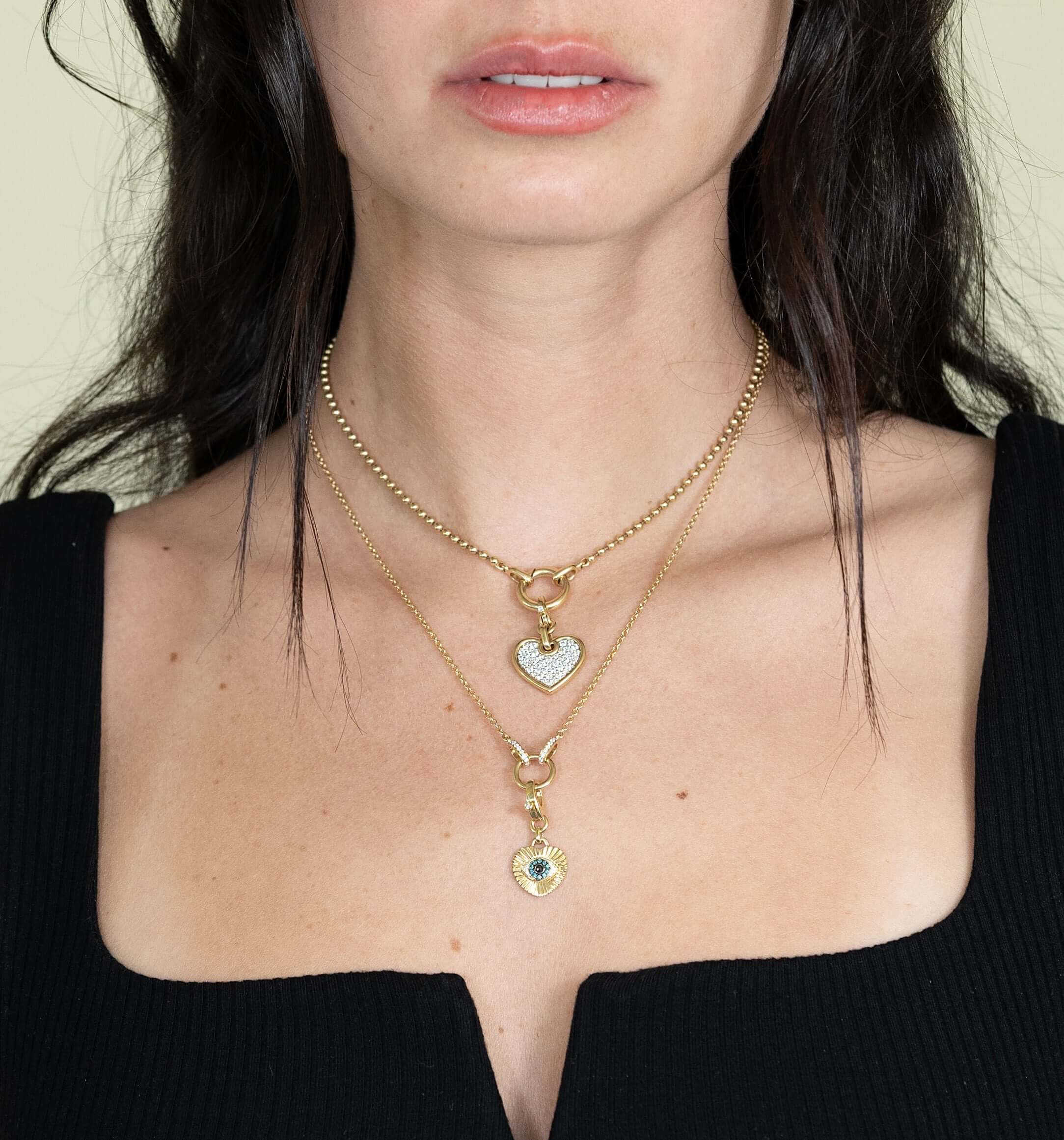 A woman wears two gold chain necklaces from Janet Heller Fine Jewelry, featuring heart-shaped pendants. The Ribbed Diamond Heart Evil Eye Charm dazzles with small stones as the upper pendant, while the lower charm has a central stone accent. She pairs them with a black square-neck top.