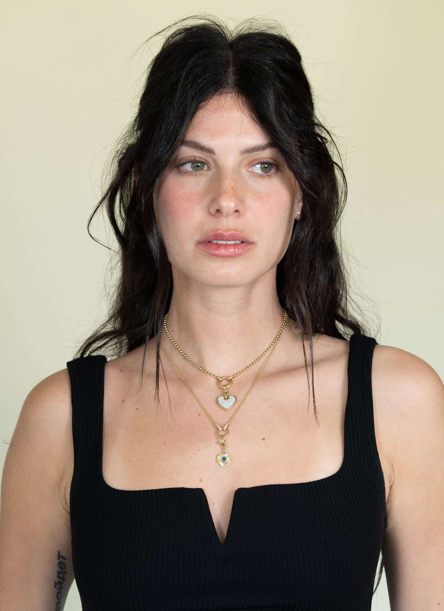 A woman with long dark hair wears a black top and layered necklaces featuring Janet Heller Fine Jewelrys Amsterdam Chain. The delicate chains glisten subtly as she looks to the side against a light-colored background.