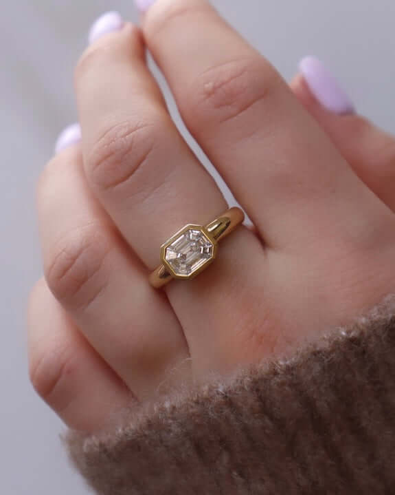 A close-up showcases a hand wearing the Diamond Athena Ring, featuring a large clear gemstone in a geometric design. The pastel-colored nails and cozy brown knit sweater complete the elegant look.