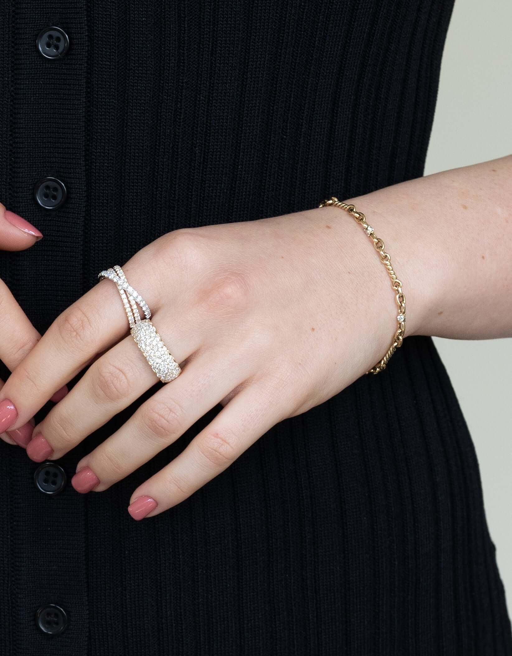 Wearing a black ribbed shirt with black buttons, a person showcases fine jewelry, including multiple sparkling rings like the Diamond Mosaic Ring and a delicate gold bracelet. Their light pink nail polish adds an elegant touch to this ensemble available for preorder.
