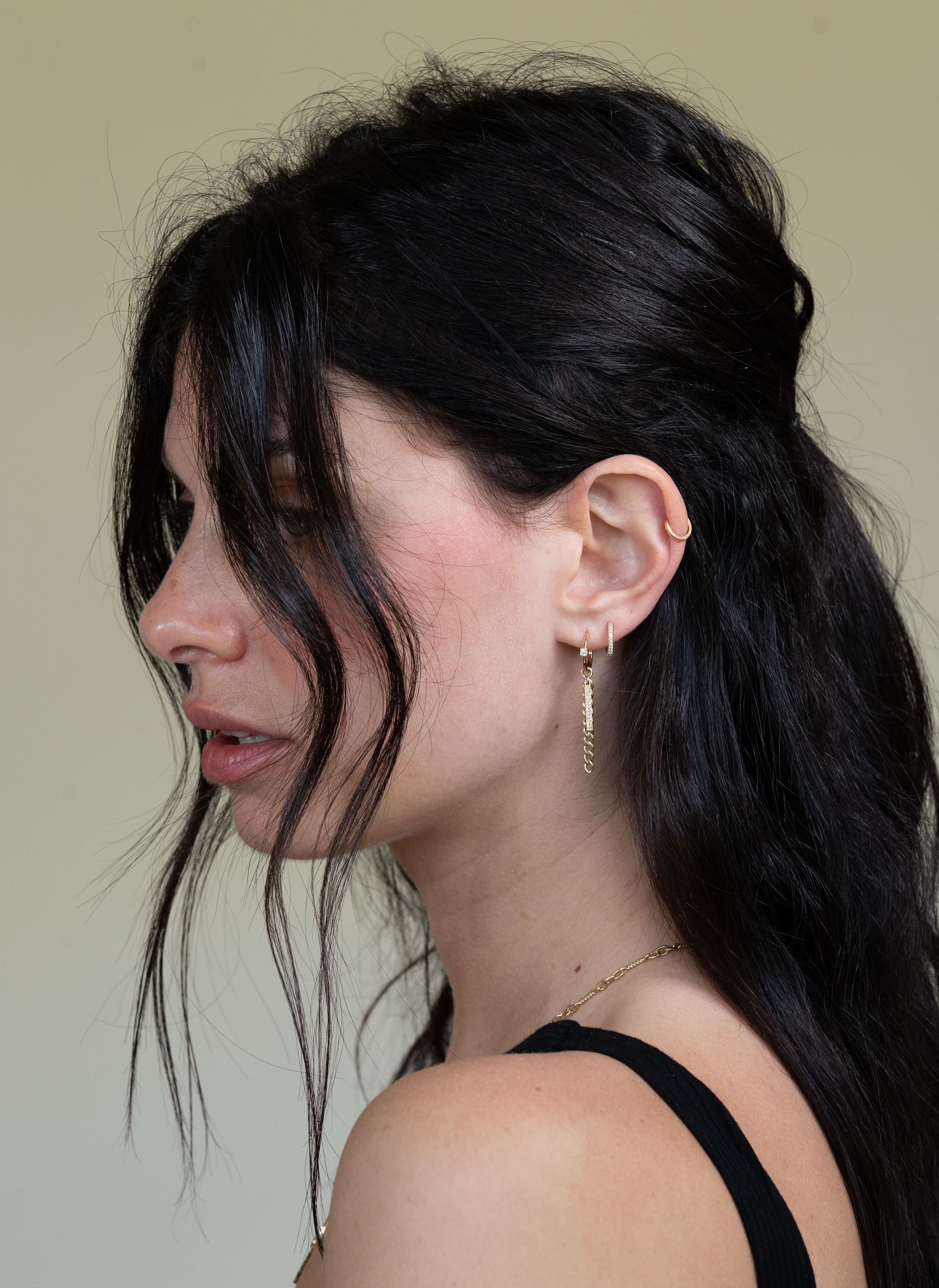 A woman with long dark hair in loose waves, adorned with Janet Heller Fine Jewelrys Diamond Curb Link Bar Huggies and a drop earring. She gazes to the side against a plain background, wearing a black top.