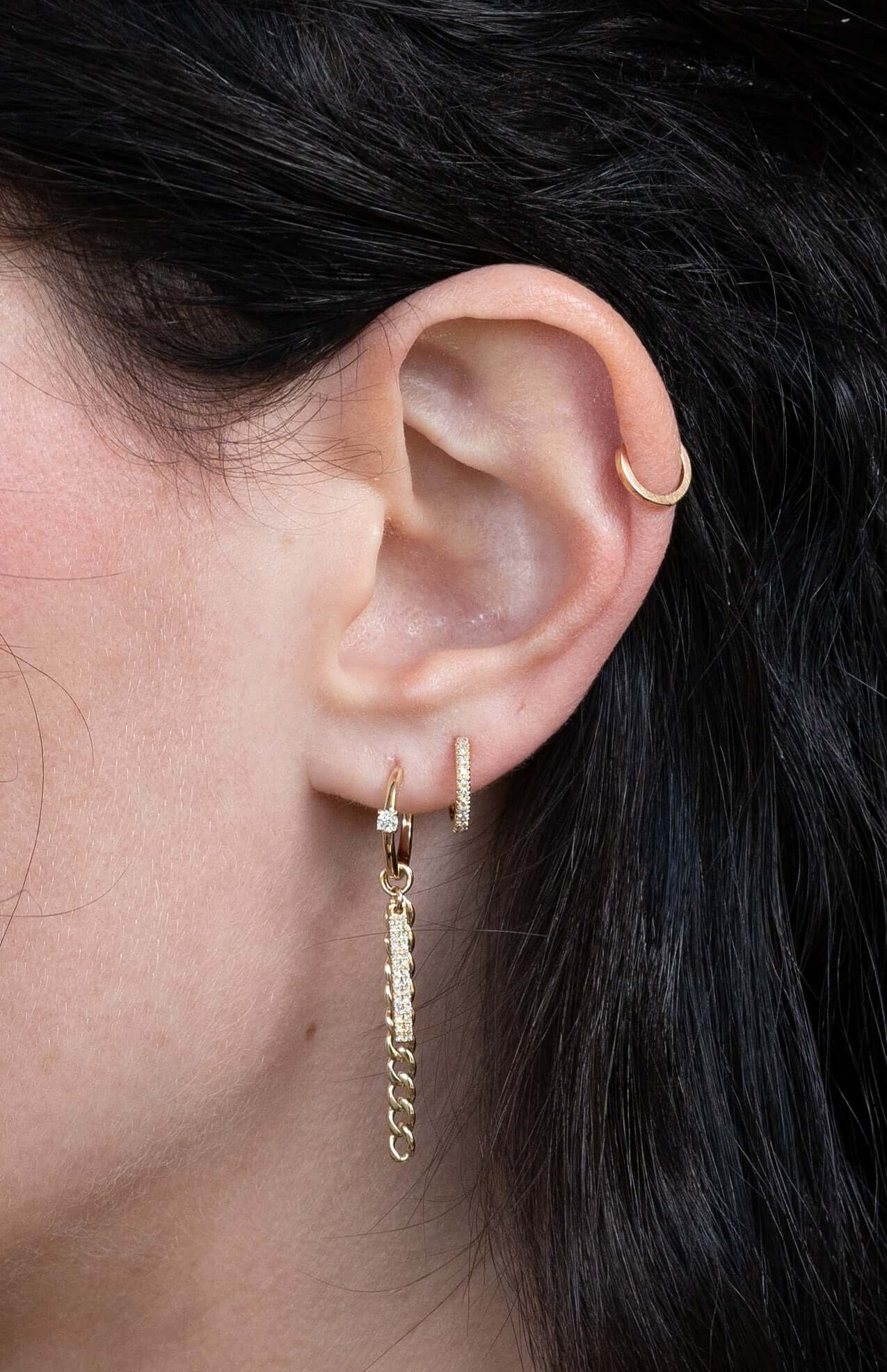 Close-up of a persons ear in janethellerfinejewelry: a small hoop on the upper ear, Diamond Curb Link Bar Huggies in the middle, and a hoop with dangling chains at the bottom. The dark-haired individual is available for customizations.