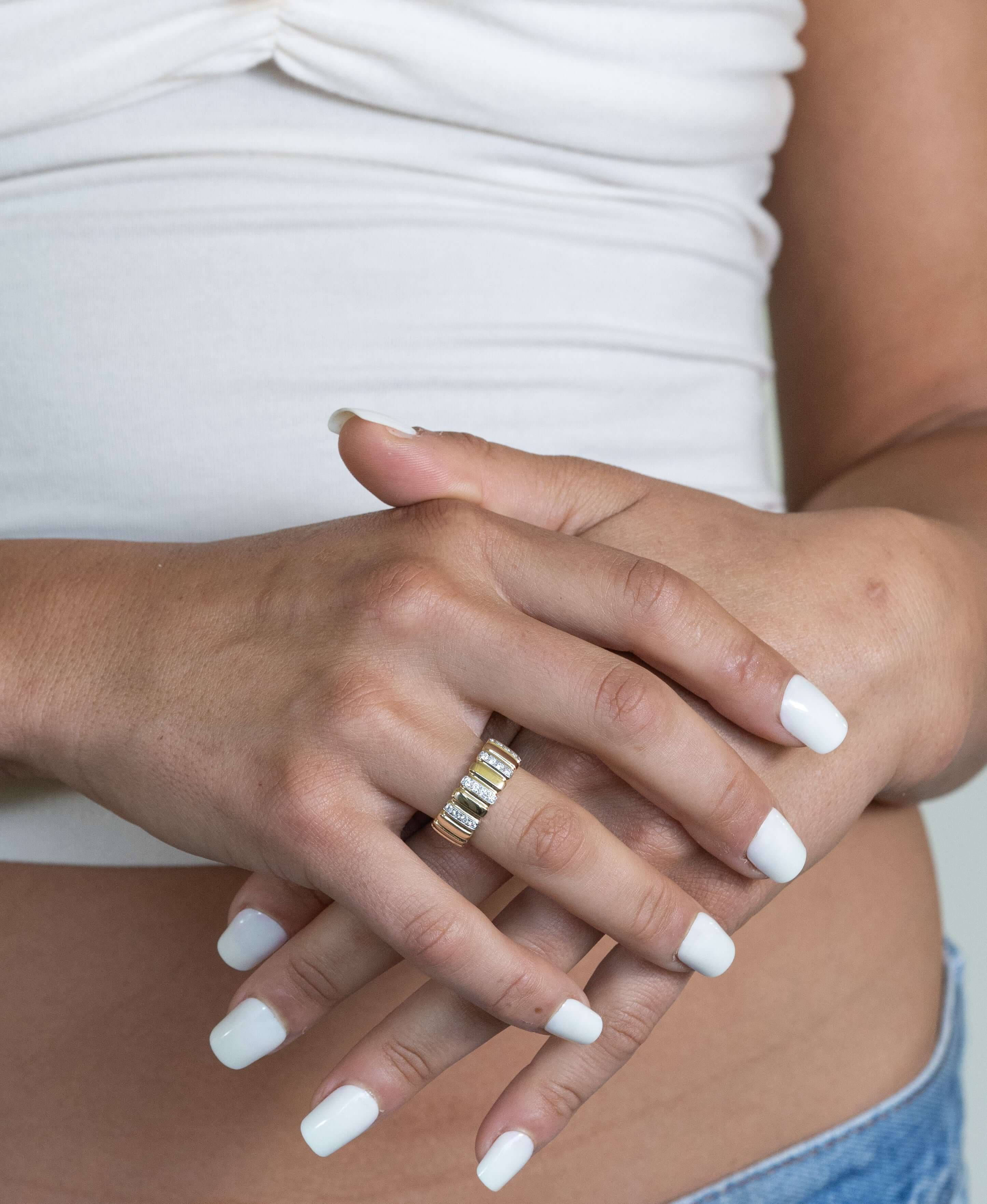 A person in a white top and jeans rests their hands, adorned with several Diamond Frankie Rings. Their nails are painted white, showcasing the jewelry beautifully against their midriff.