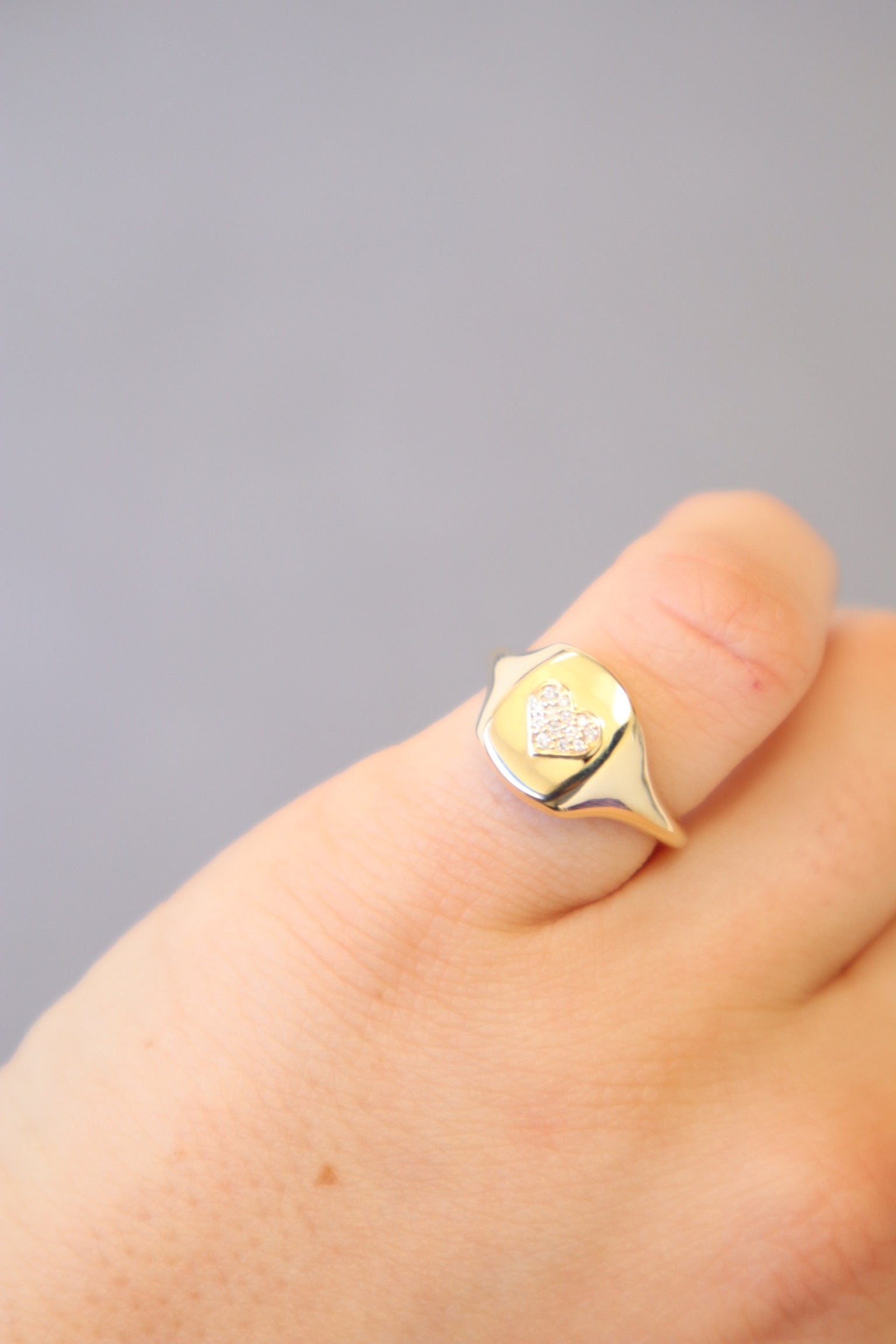 A hand wearing the Diamond Heart Signet Ring, featuring a small heart design adorned with tiny clear stones at the center, is set against a soft gray background—this stunning ring is available for preorder from Janet Heller Fine Jewelry in your preferred size.