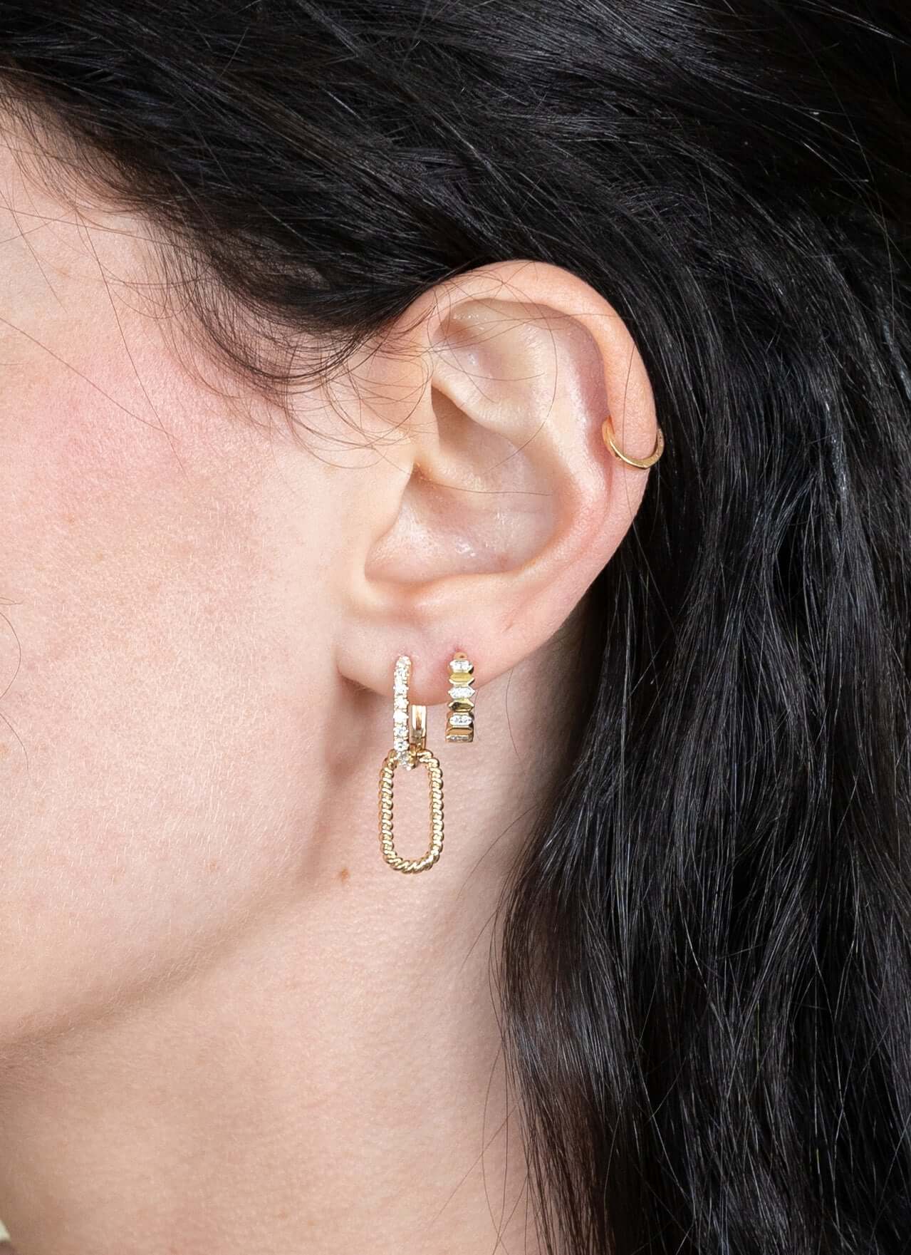 Close-up of an ear wearing custom jewelry, featuring a gold hoop on the helix and a lobule adorned with chain-link earrings, stacked gold bars, and a star stud with stones. Also visible is the Diamond Shape Gigi Huggies earring set against dark hair.