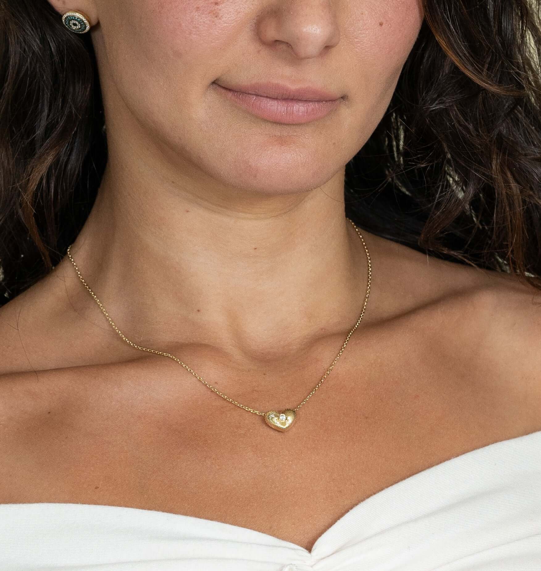 A woman wears a Diamond Puff Heart Ribbed Necklace and black earrings. Her shoulders are draped in white fabric, showcasing the stunning necklace available for customization to create your perfect product.