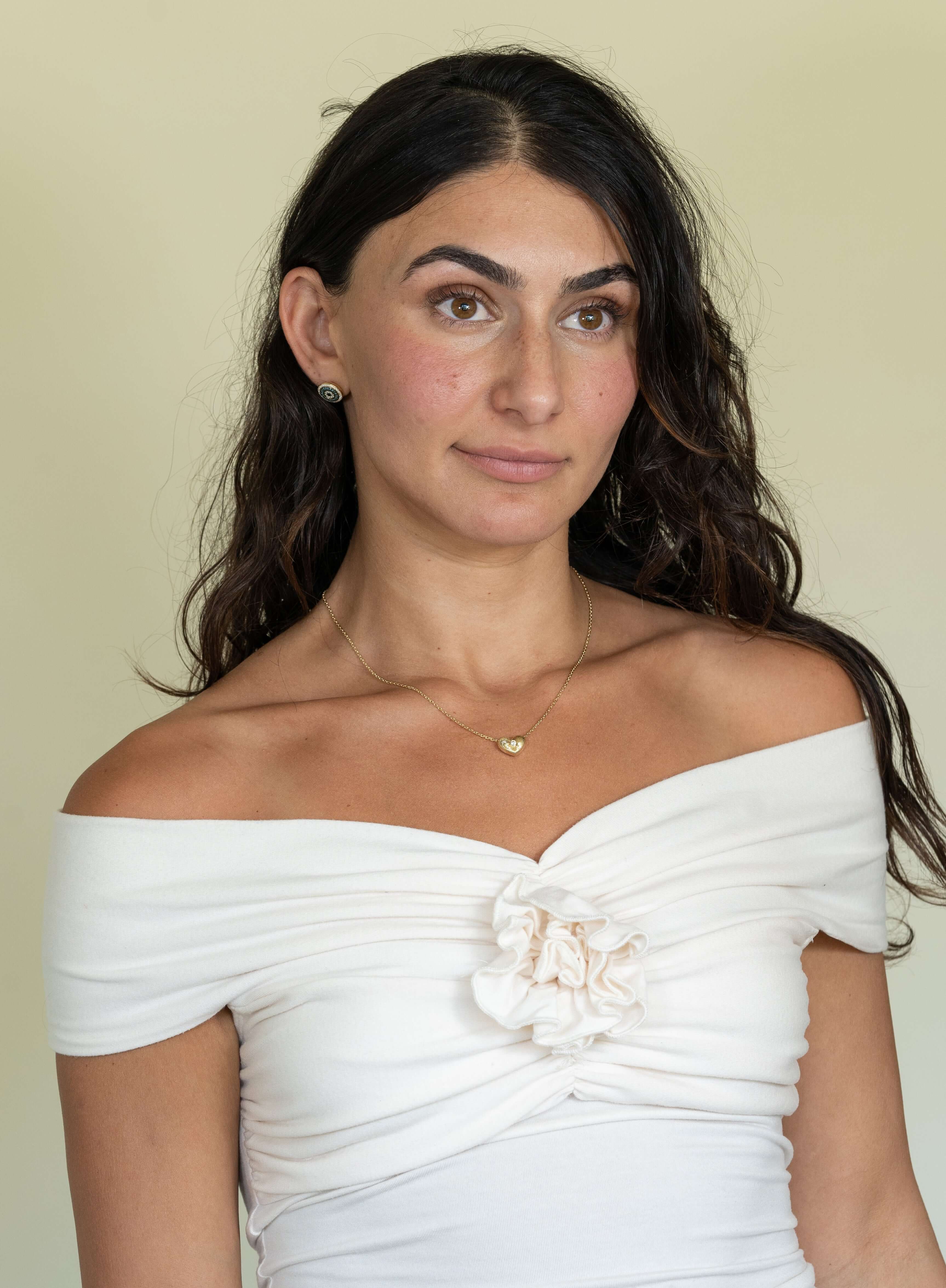 Against a plain beige background, a person with long dark hair and a warm complexion wears an off-shoulder white top with floral detail. Their look is enhanced by custom jewelry, including small hoop earrings and the Diamond Puff Heart Ribbed Necklace.
