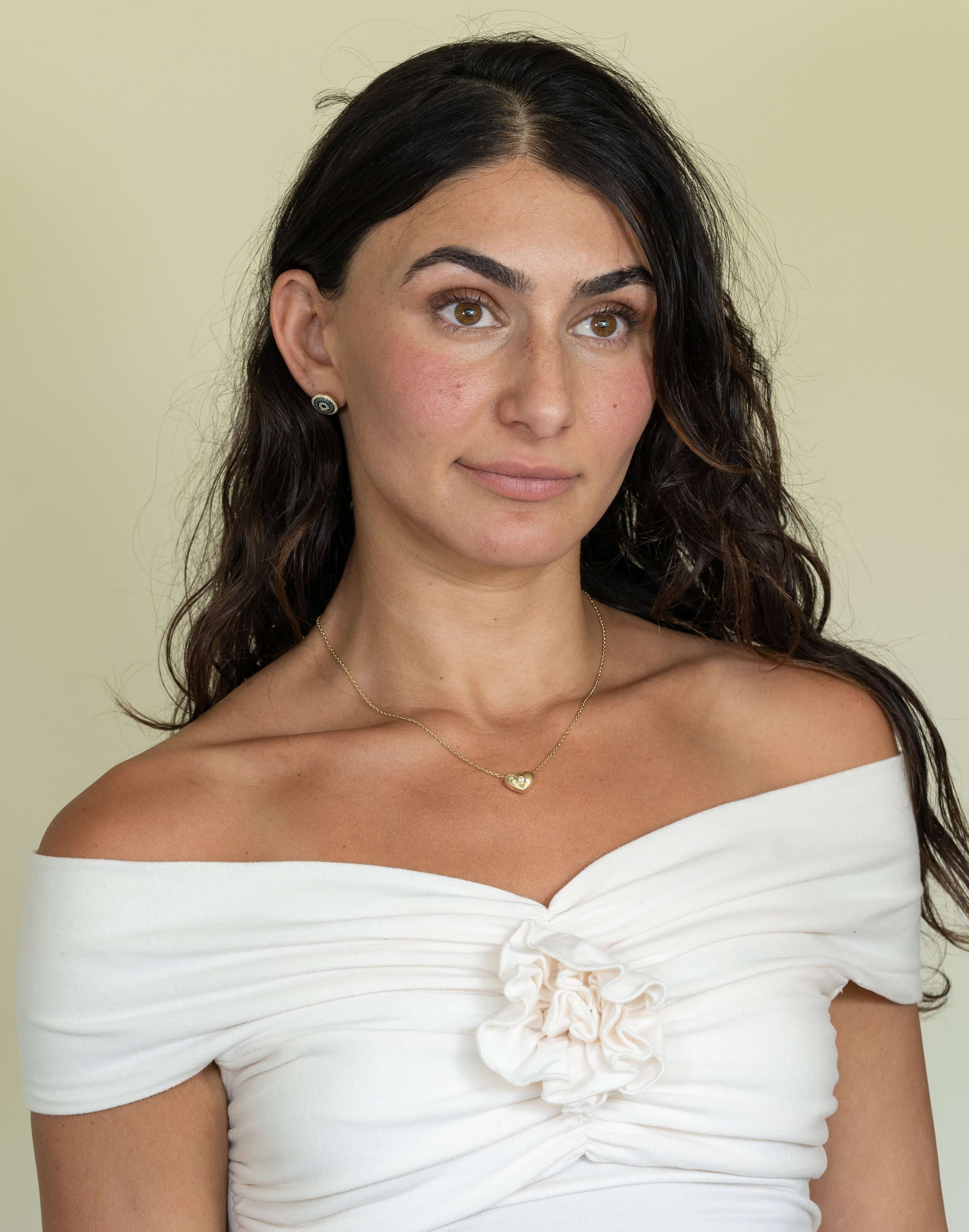 A woman with long, dark hair in a white floral off-shoulder top stands against a beige background. She elegantly wears the Diamond Puff Heart Ribbed Necklace and small stud earrings.