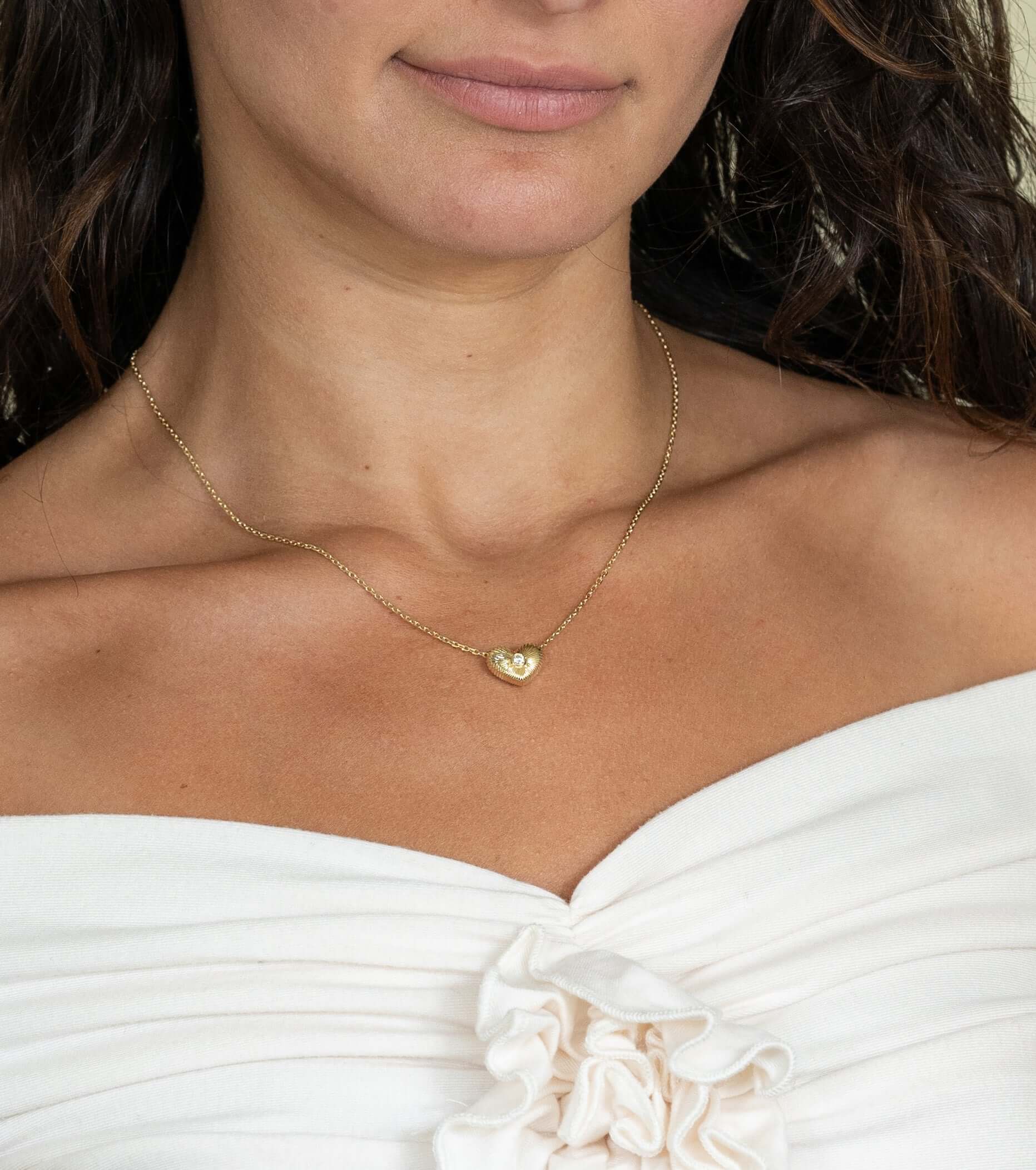 A woman in an off-the-shoulder white ruffled top effortlessly showcases her jewelry. Her dark wavy hair frames a delicate gold chain, the Diamond Puff Heart Ribbed Necklace, with a heart-shaped pendant resting just below her collarbone.