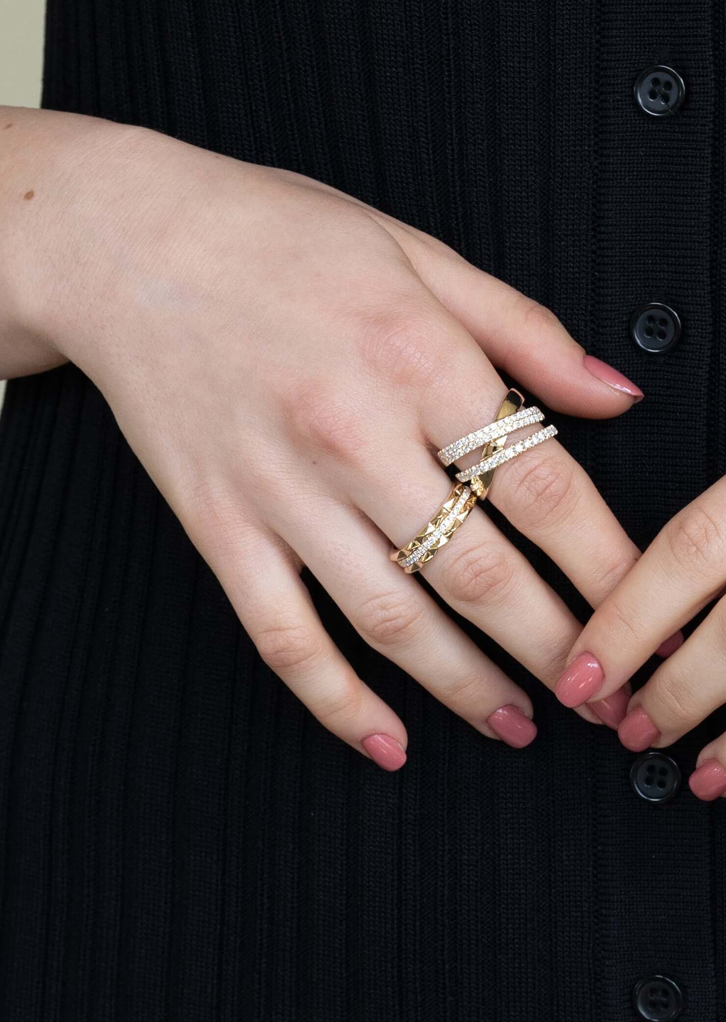 A persons hand with pink nail polish rests on black ribbed fabric, showcasing exquisite rings by Janet Heller Fine Jewelry. The centerpiece is the Diamond Pyramid Triple Row Ring. Multiple gold and silver bands with small gems grace the fingers. Preorder your favorite piece today.