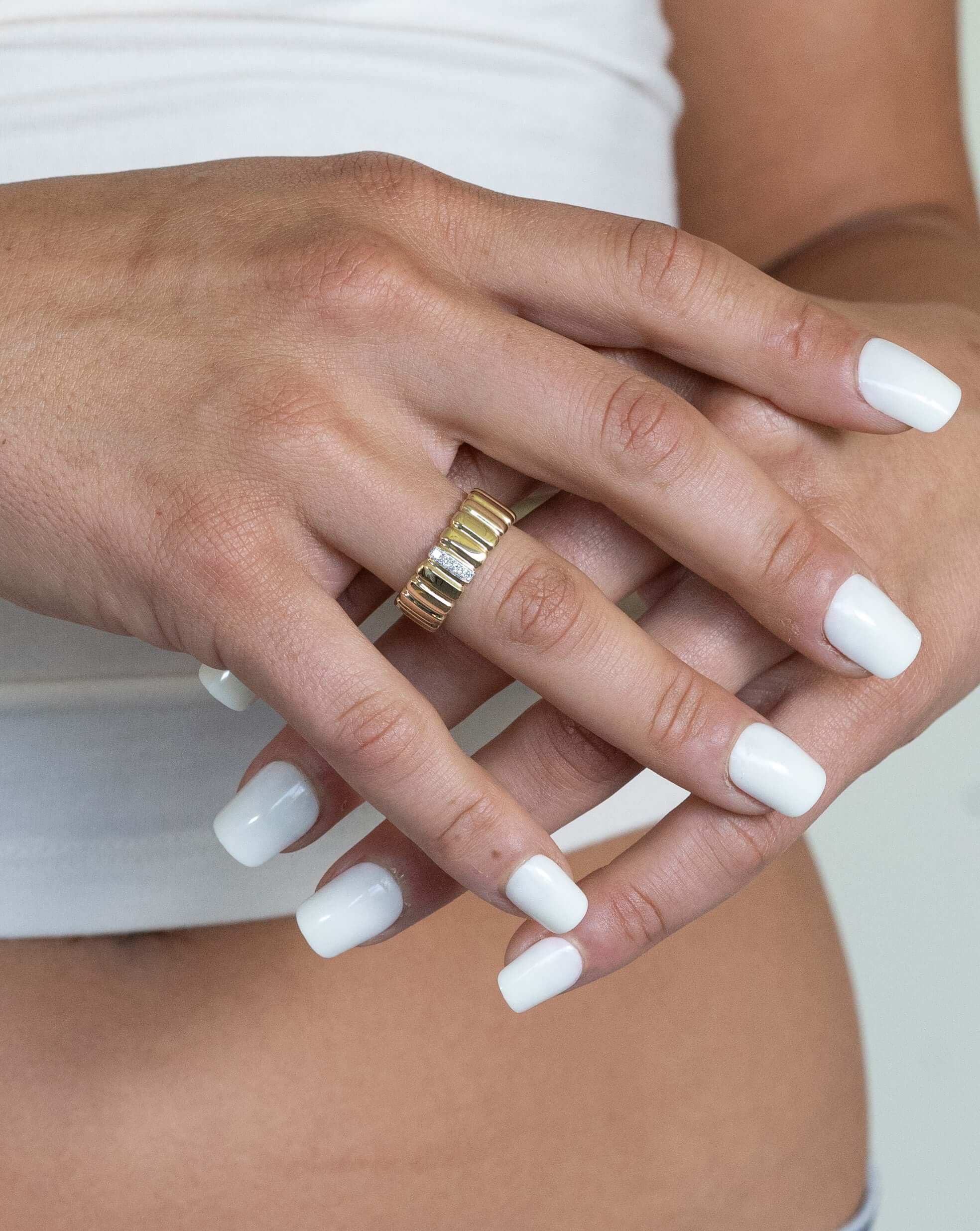 Close-up of a person with neatly manicured white nails, wearing the preorder Diamond Ridge Rio Ring with multiple bands. Their hand rests gently over the other against a light background, showcasing fine jewelry while wearing a white top.