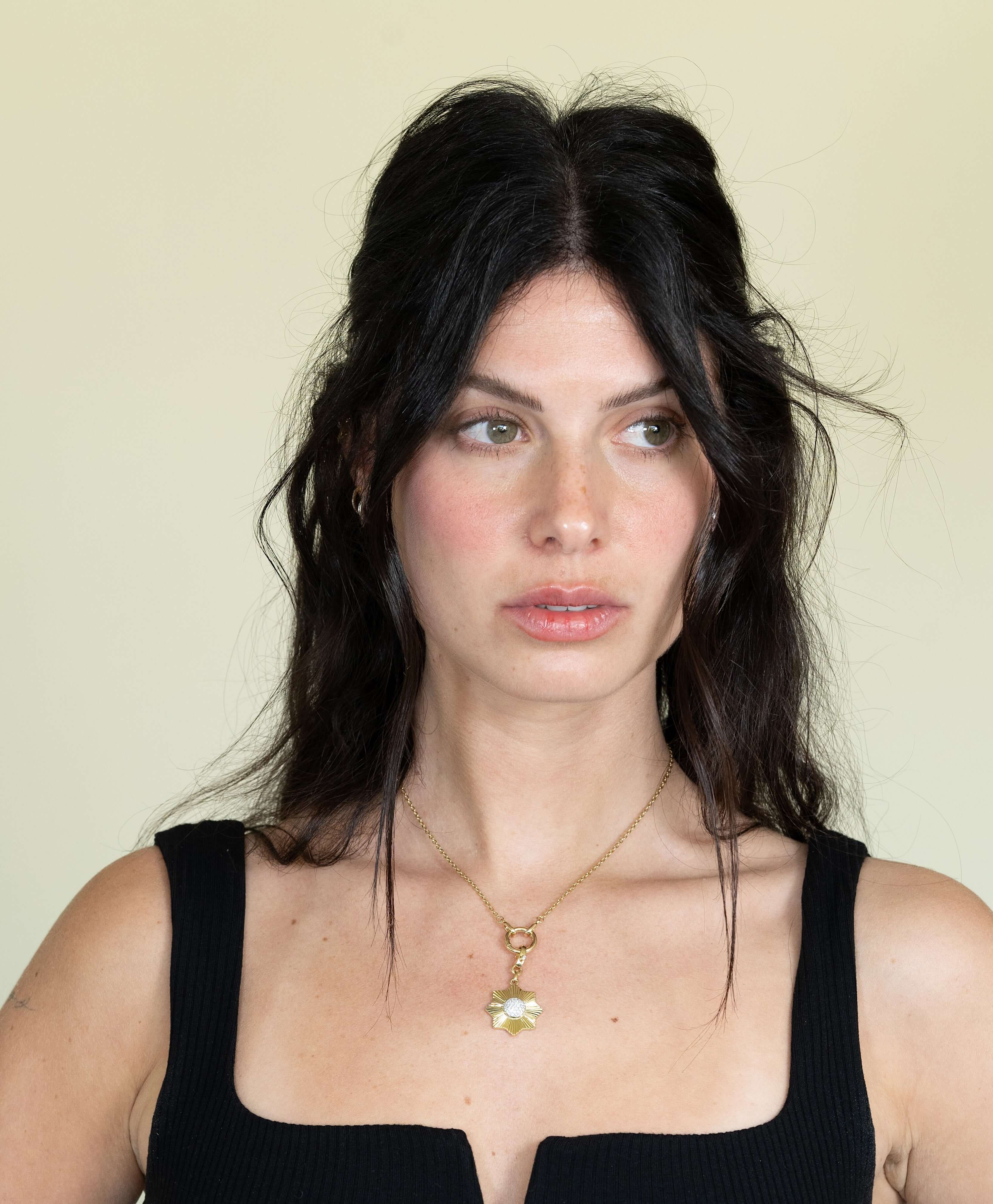 A woman with long dark hair styled back, wearing a sleeveless black top and a gold necklace featuring the Pave Diamond Sun Flower Charm, gazes slightly to the side against a plain background.