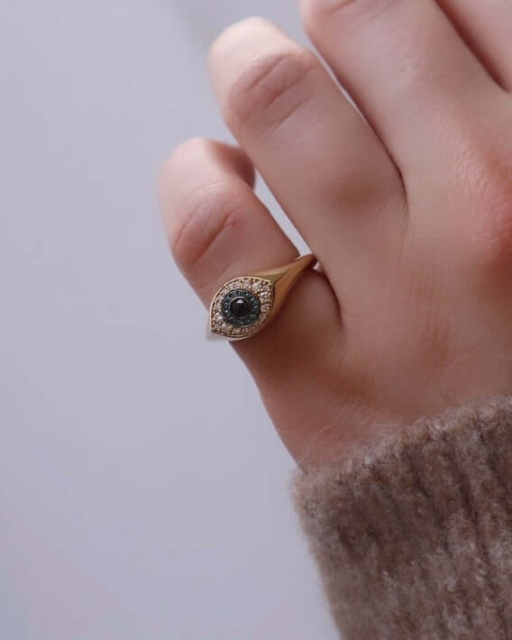 The Evil Eye Pinkie Ring by Janet Heller Fine Jewelry is crafted in 14KT gold. The ring features multi-colored diamonds in white and blue with a rose-cut black center diamond.
