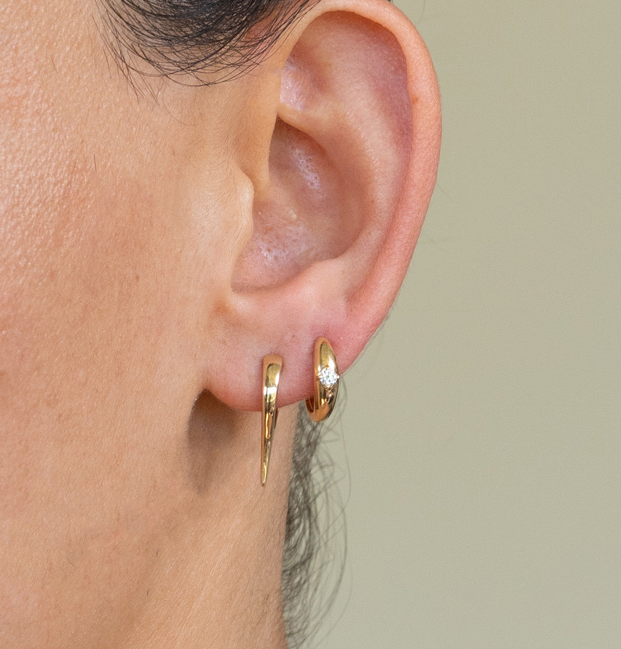 Close-up of a persons ear adorned with Janet Hellers Solitaire Diamond Dome Huggies: a small gold hoop with a tiny gem on the earlobe and an elongated hoop in the lower piercing. The background is soft and neutral.