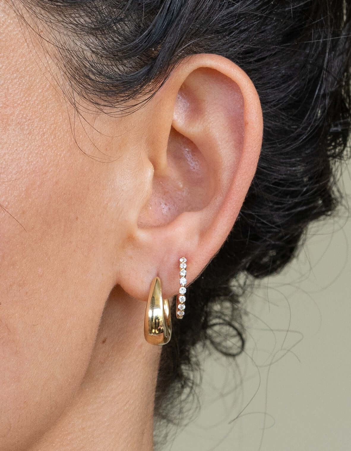 A close-up shows a person wearing earrings from Janet Heller Fine Jewelry: a thick, shiny gold Oval Dome Hoop on the lower lobe and a slender curved bar with small white stones above it. Inquiries or customizations are welcome. Neutral background.