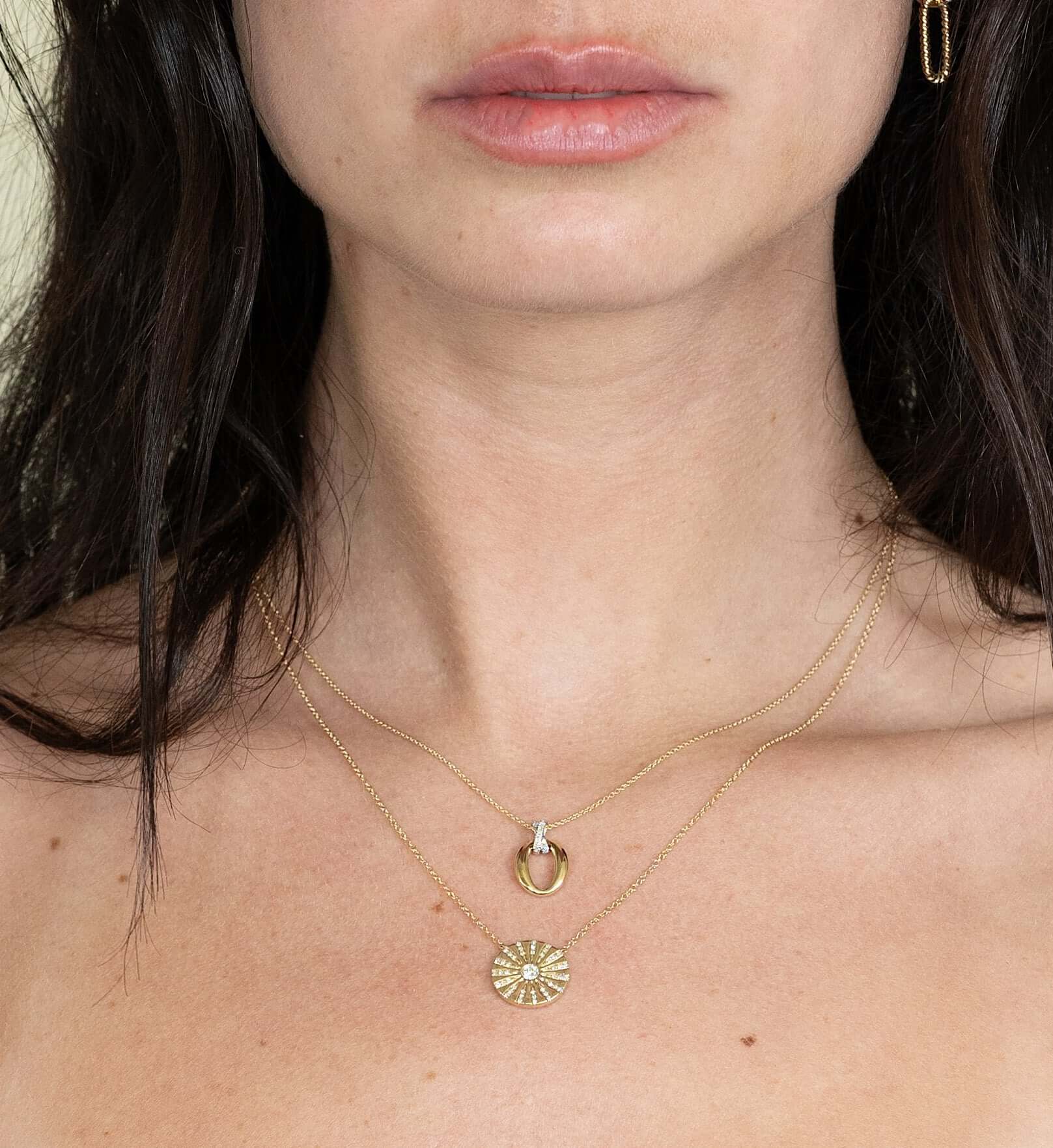A woman with long dark hair wears two delicate gold necklaces from janethellerfinejewelry, including the Del Sol Diamond Circle Necklace with its starburst pendant, and another featuring a small ring. She complements her look with a subtle gold earring, highlighting exquisite customizations.