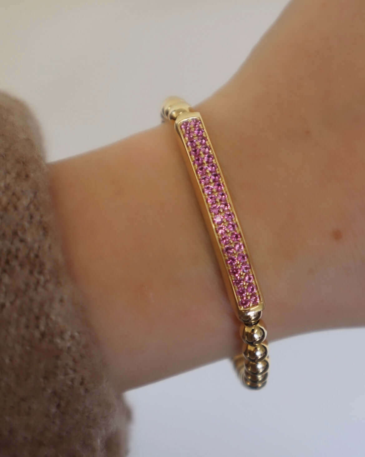 A Janet Heller Fine Jewelry creation, the Pink Sapphire Lennon Stretchy Bracelet, features a gold rectangular bar with small pink stones. Crafted from round, linked beads, it elegantly rests on a wrist against a brown sleeve.