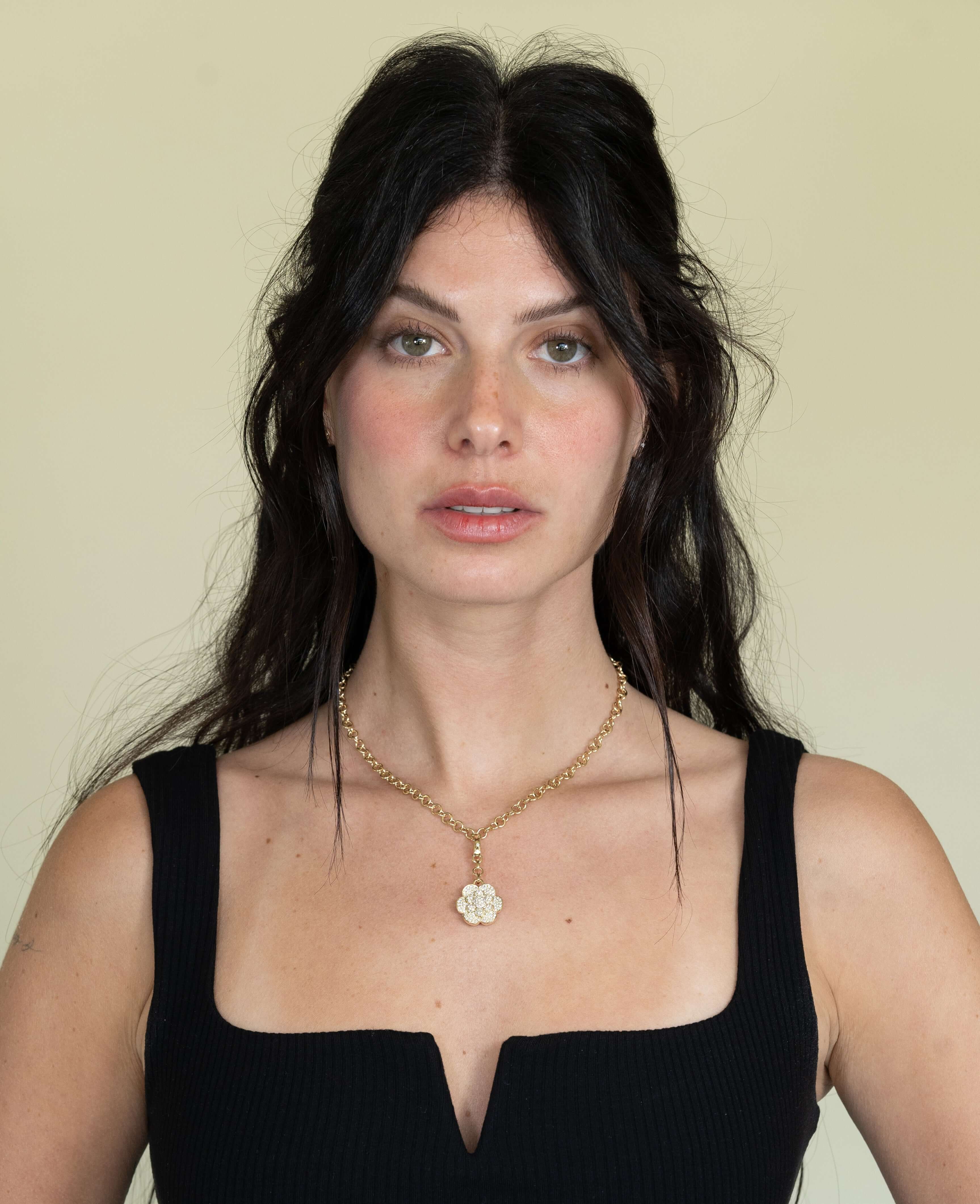 A person with long dark hair is wearing a black top and necklace featuring the exquisite Diamond Bloom Flower Charm by Janet Heller Fine Jewelry. They have a neutral expression and are looking directly at the camera against a plain background.