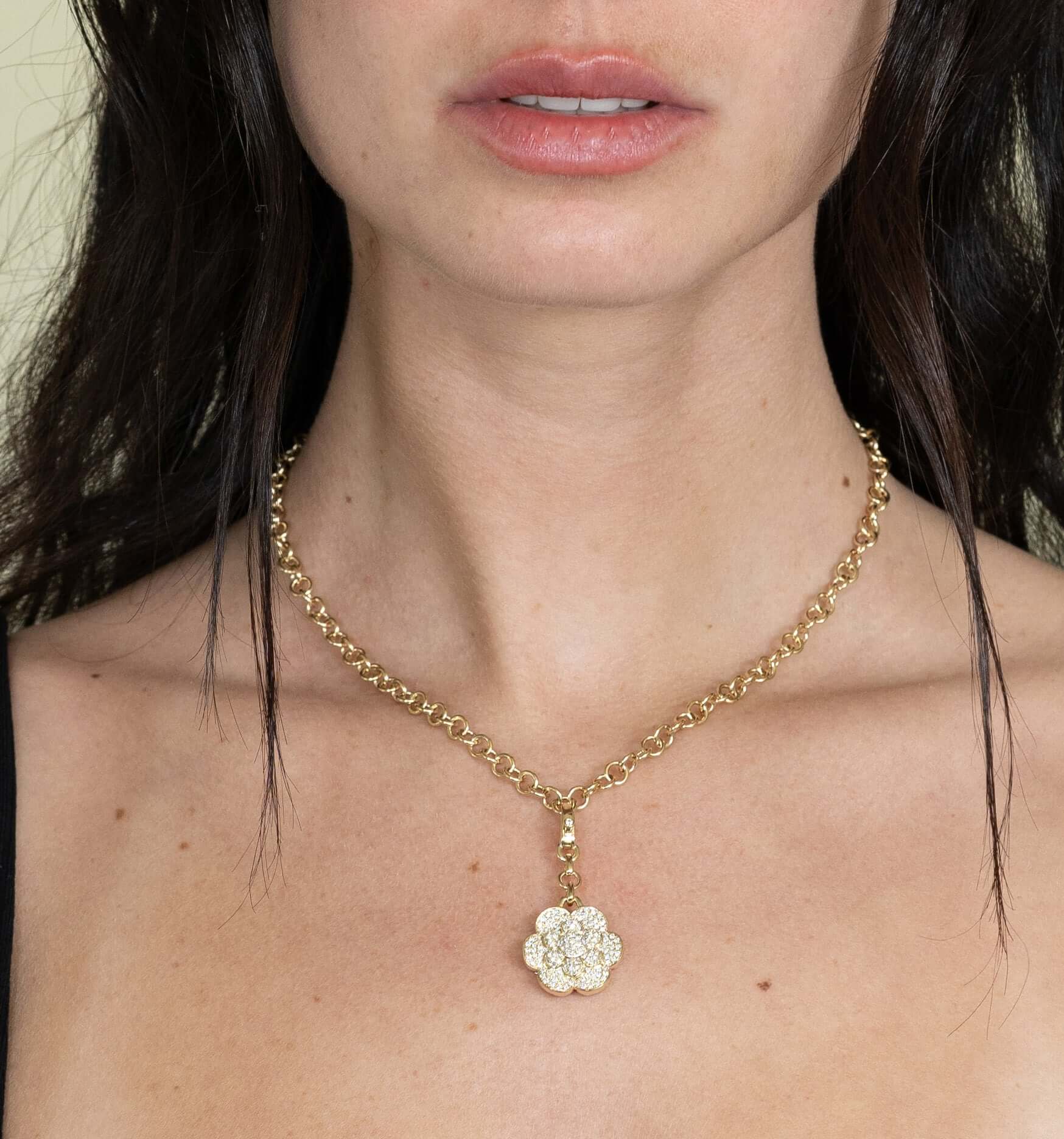 A woman with dark hair wears the Diamond Bloom Flower Charm from Janet Heller Fine Jewelry, a gold chain necklace with a gemstone-adorned flower pendant. The image elegantly showcases the necklace and her lower face, highlighting subtle customizations.