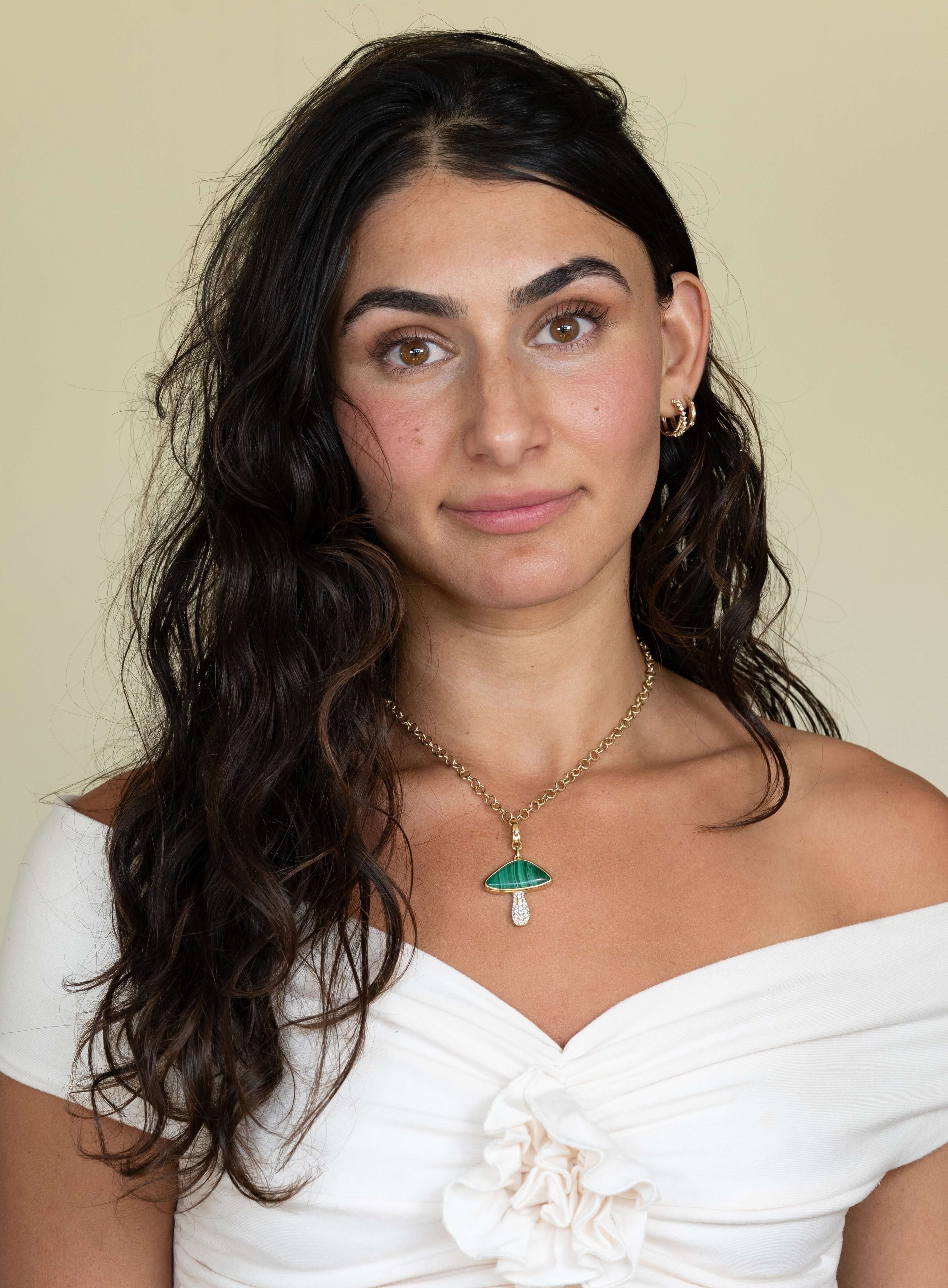 A woman with long, wavy dark hair wears a white off-the-shoulder top and stares at the camera. Her gold hoop earrings and the Round Link Phoenix Chain with a mushroom-shaped pendant reflect Janet Heller Fine Jewelrys elegance against a neutral beige background.