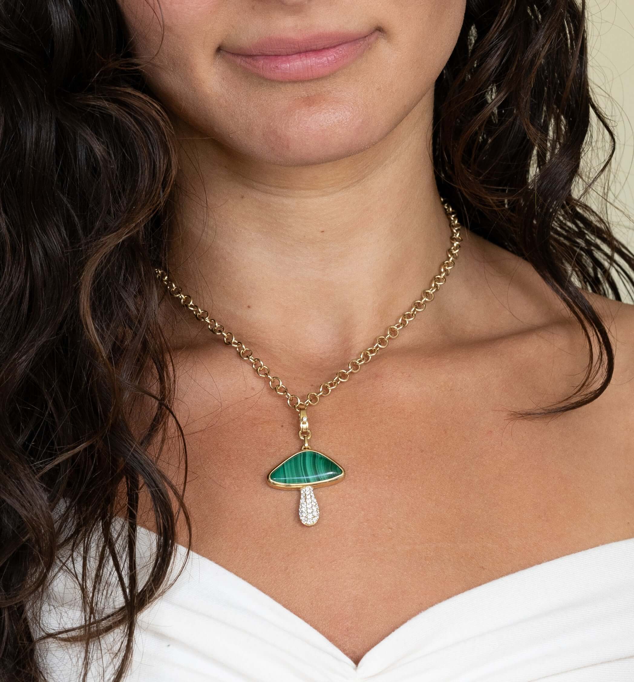 A person with long, wavy hair wears the Round Link Phoenix Chain from Janet Heller Fine Jewelry, featuring a pendant shaped like a green mushroom with white details. They pair it with an off-shoulder white top.