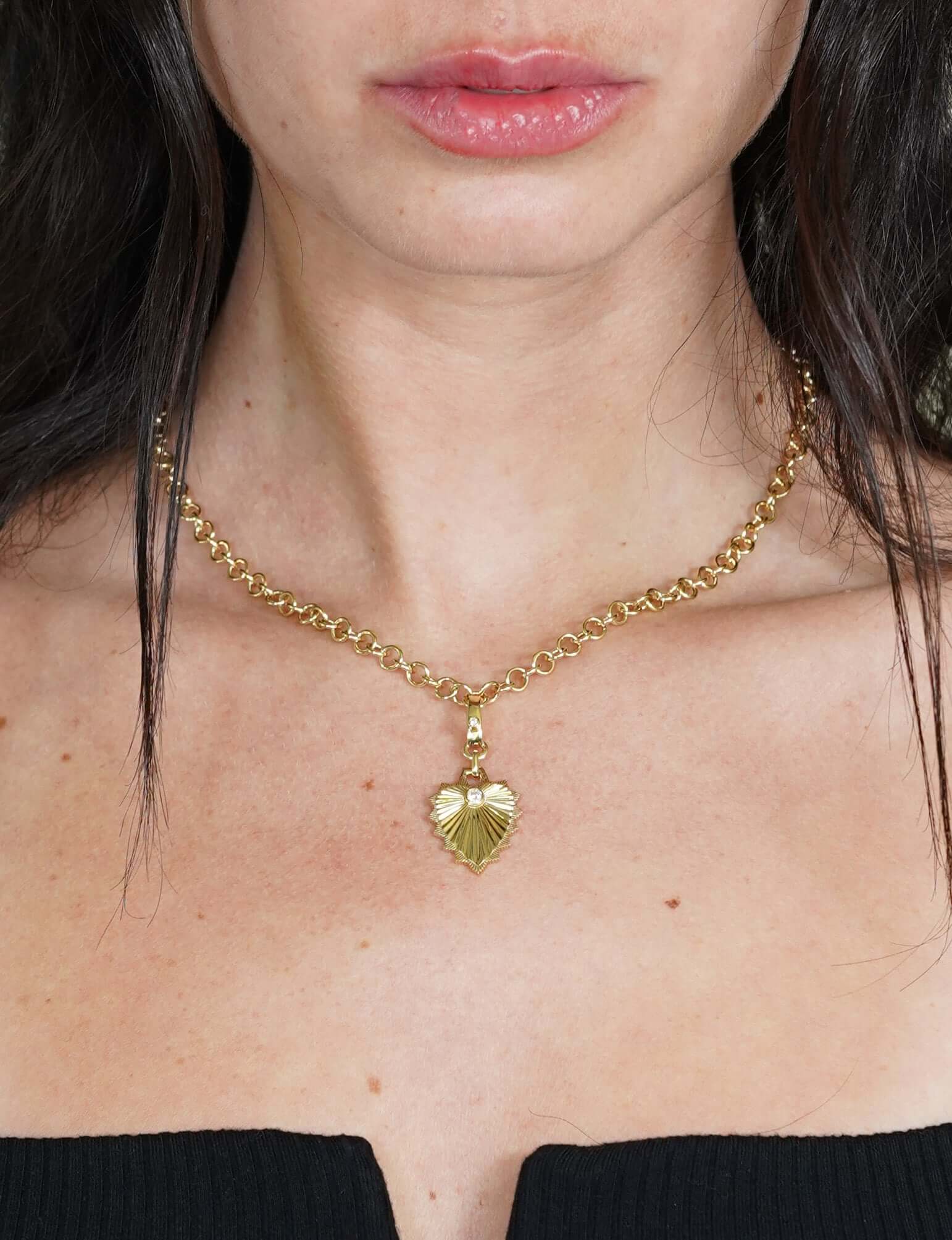 A person wearing a Round Link Phoenix Chain with a heart-shaped pendant and pearl-like accents from Janet Heller Fine Jewelry. Their long dark hair enhances a chic black, off-the-shoulder top. For inquiries or customizations, contact us.