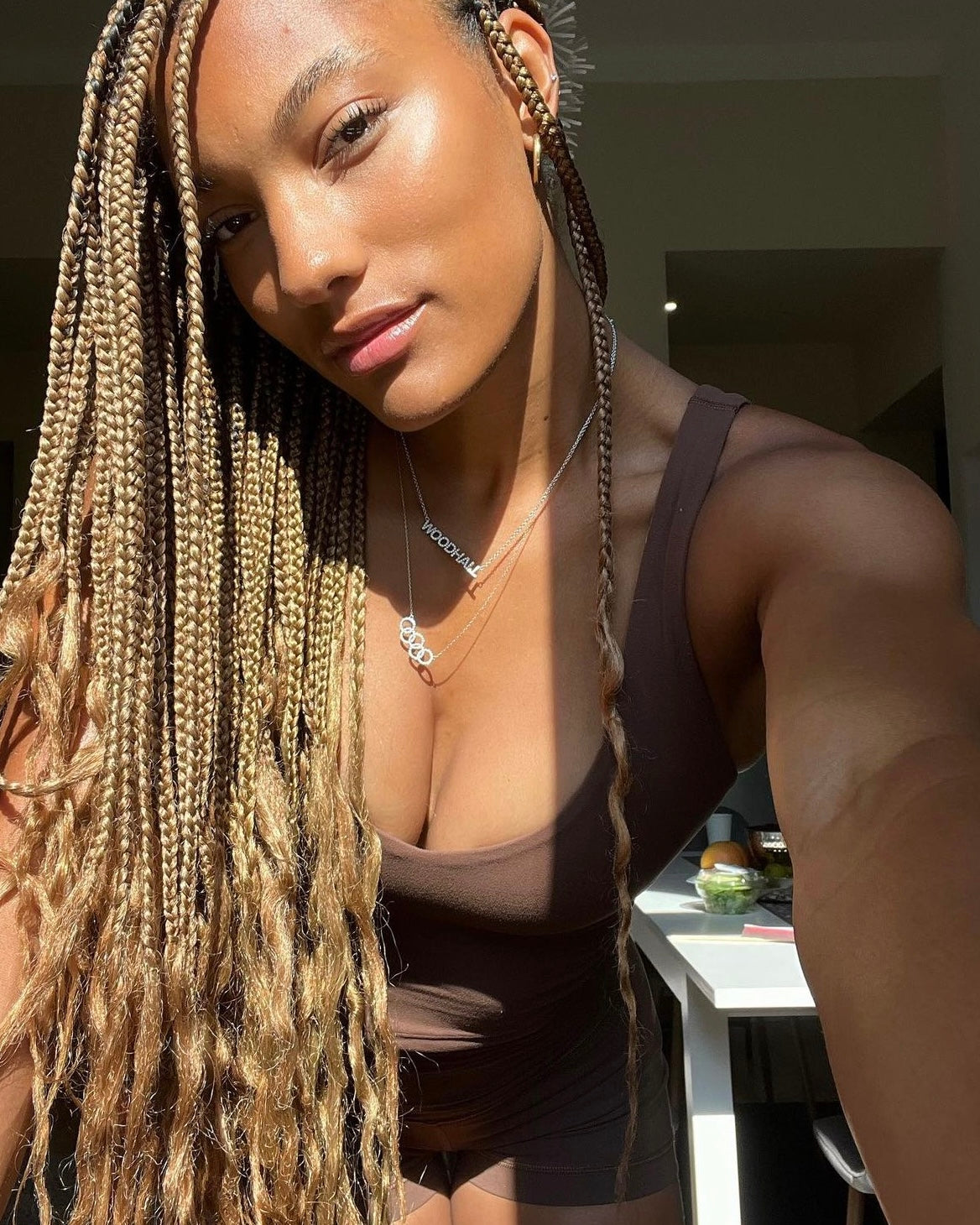 A woman with long, blonde box braids takes a selfie in sunlight. She wears a brown tank top and the Diamond Rings Necklace, gazing softly into the camera. A table with pre-order items appears in the background.