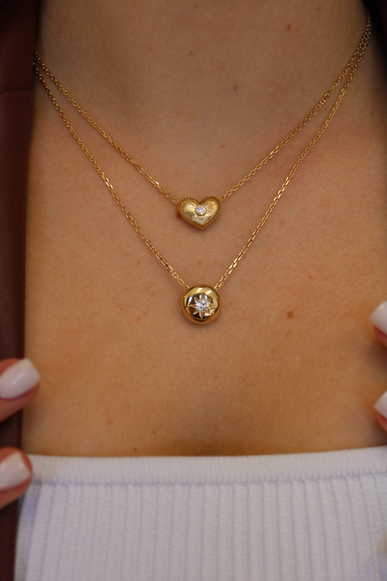 A person showcases exquisite jewelry, with two gold necklaces on display. The top is the Diamond Puff Heart Ribbed Necklace featuring a heart-shaped pendant with a small gemstone, while the bottom necklace has a round pendant with a larger gemstone, elegantly paired with a white top.