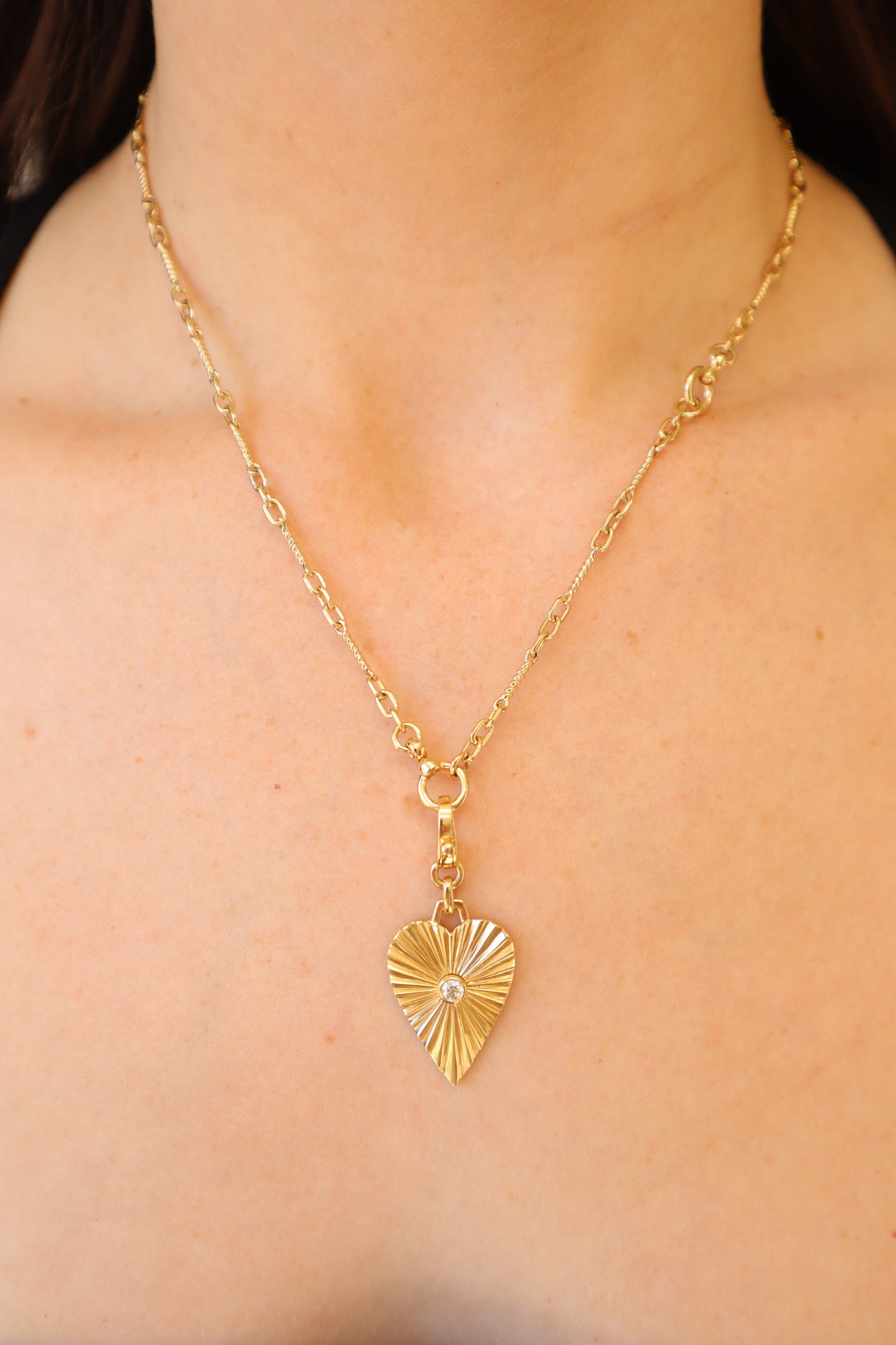 A person wears a gold chain necklace from Janet Heller Fine Jewelry, showcased by the Diamond Heart Burst Charm with its textured heart-shaped pendant and intricate center detail.